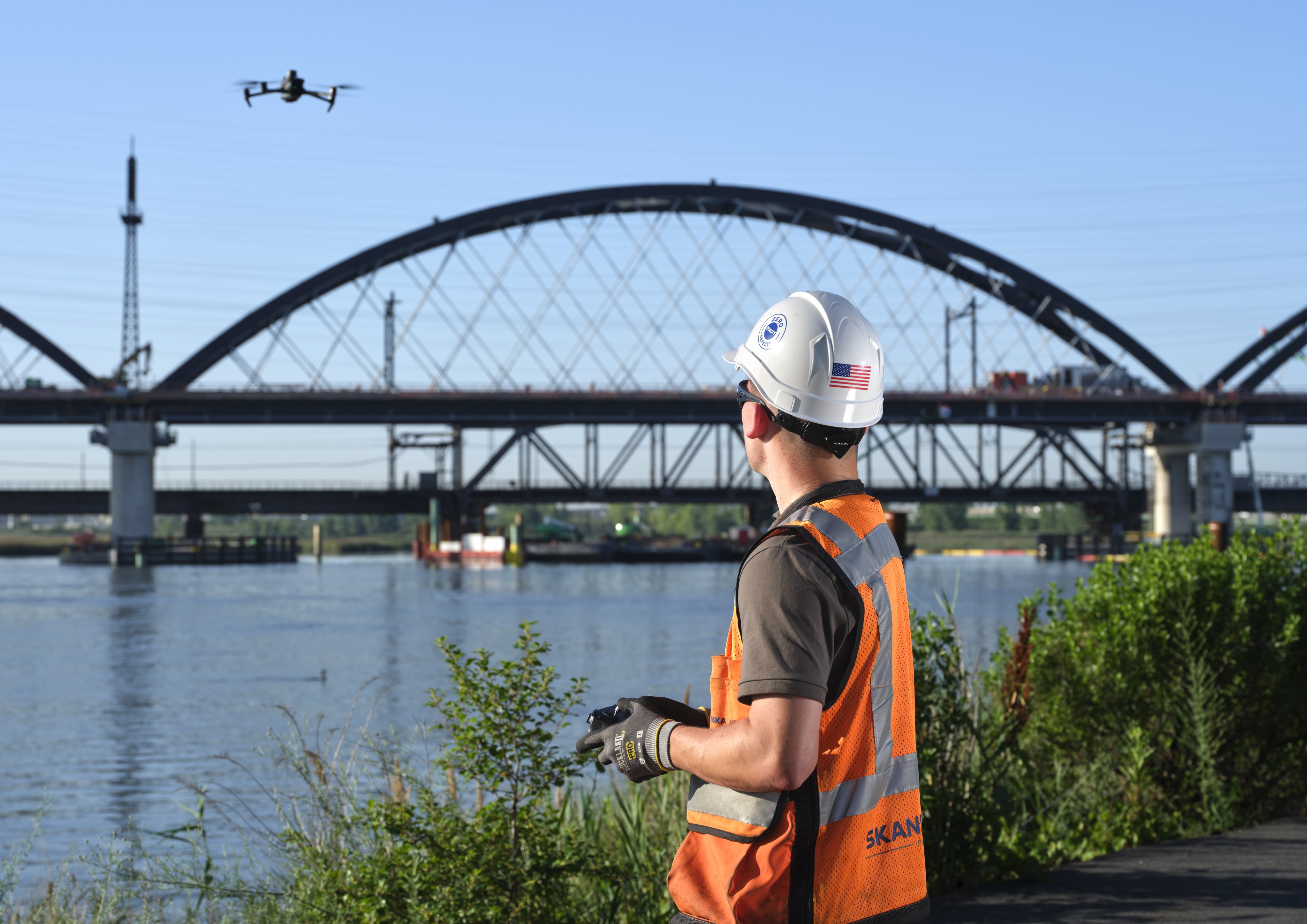 A construction worker operates a drone near a large bridge over water, wearing safety gear.