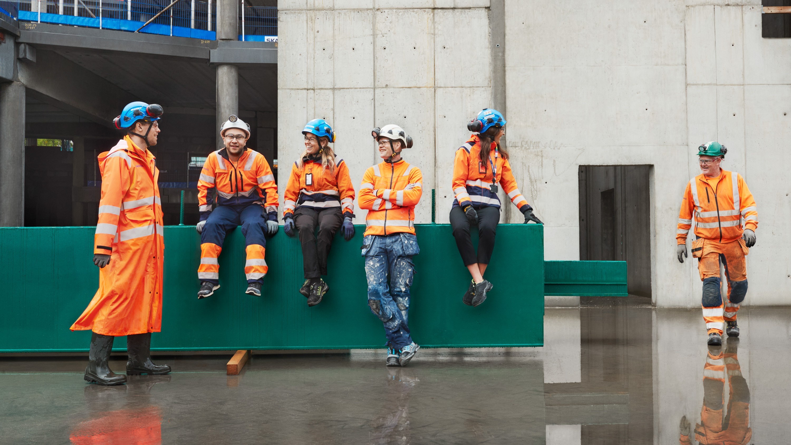 Construction workers in orange safety gear and helmets converse at a building site.