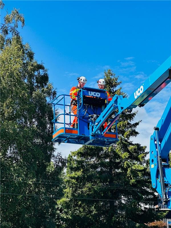 To arbeidere i verneutstyr står på en blå UCO luftlift, omgitt av trær under en klar himmel.