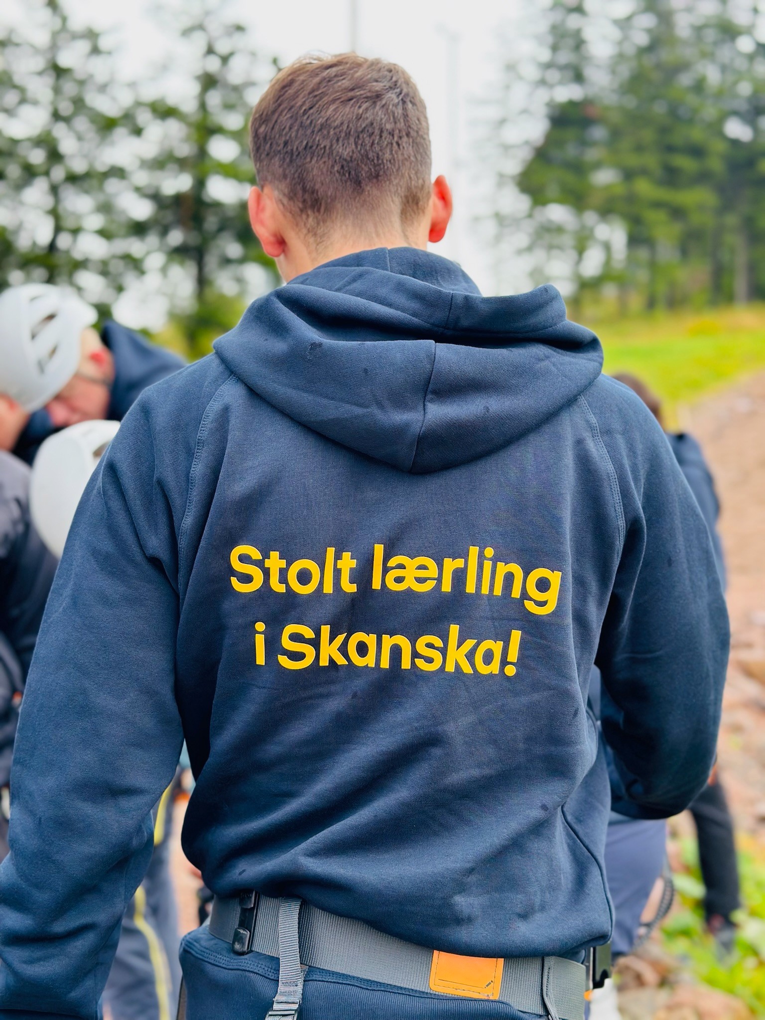 Person iført hettegenser med teksten "Stolt lærling hos Skanska!" som står utendørs med trær i nærheten.