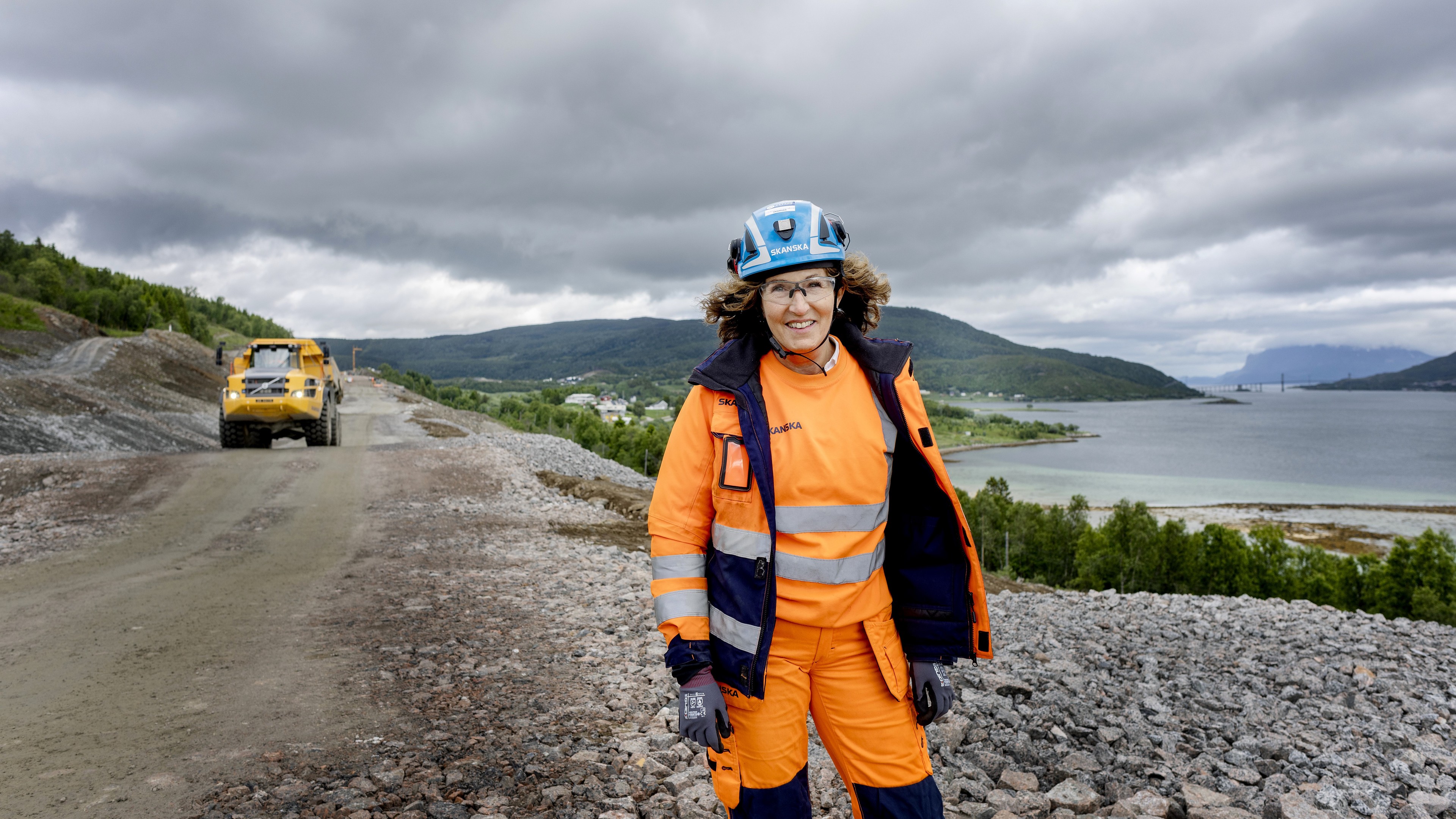 Bygningsarbeider i høy synlighetstøy står på en steinete sti nær en naturskjønn innsjø.