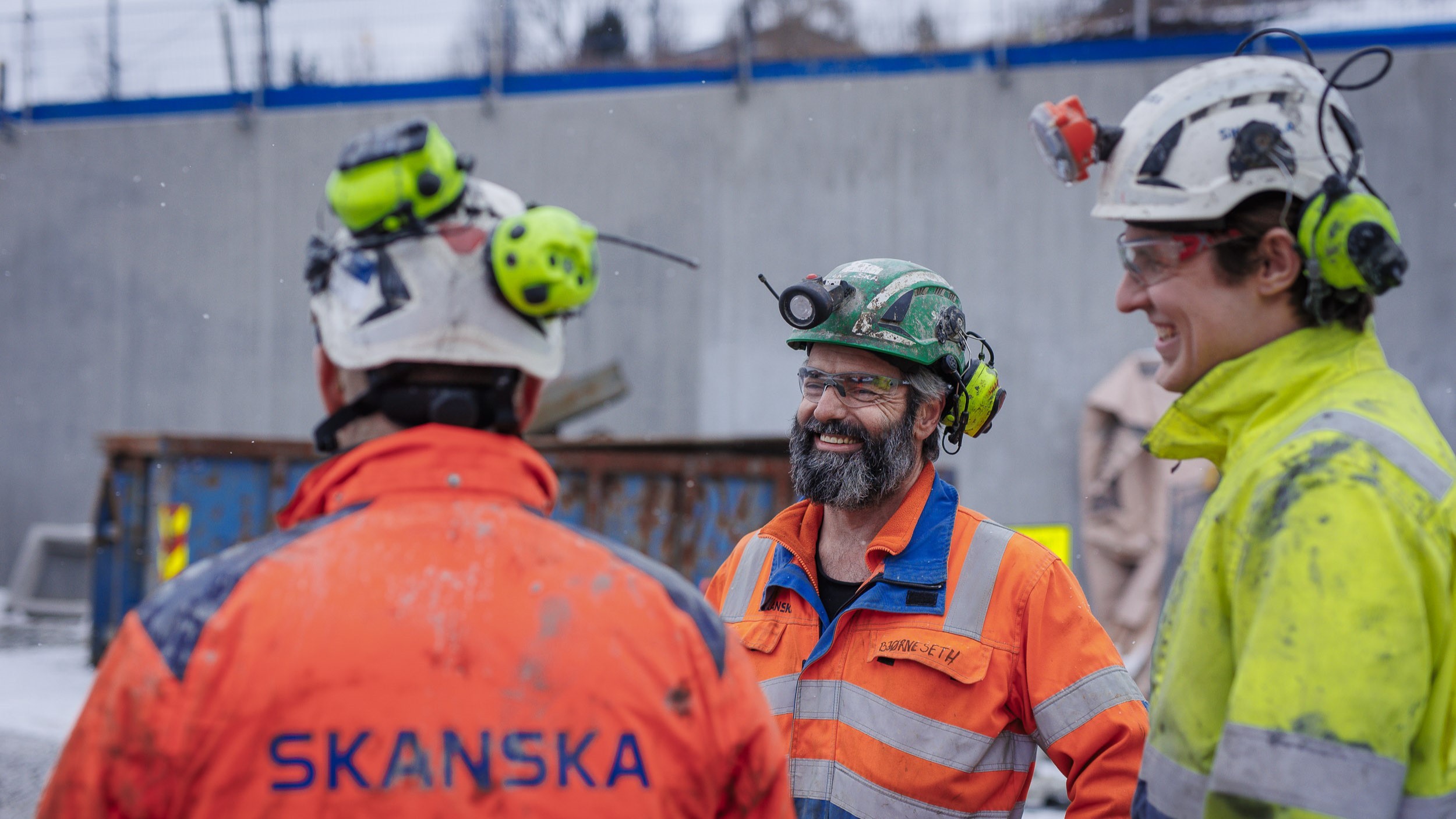 Bygningsarbeidere i verneutstyr og hjelmer på en utendørs byggeplass med Skanska-logo.