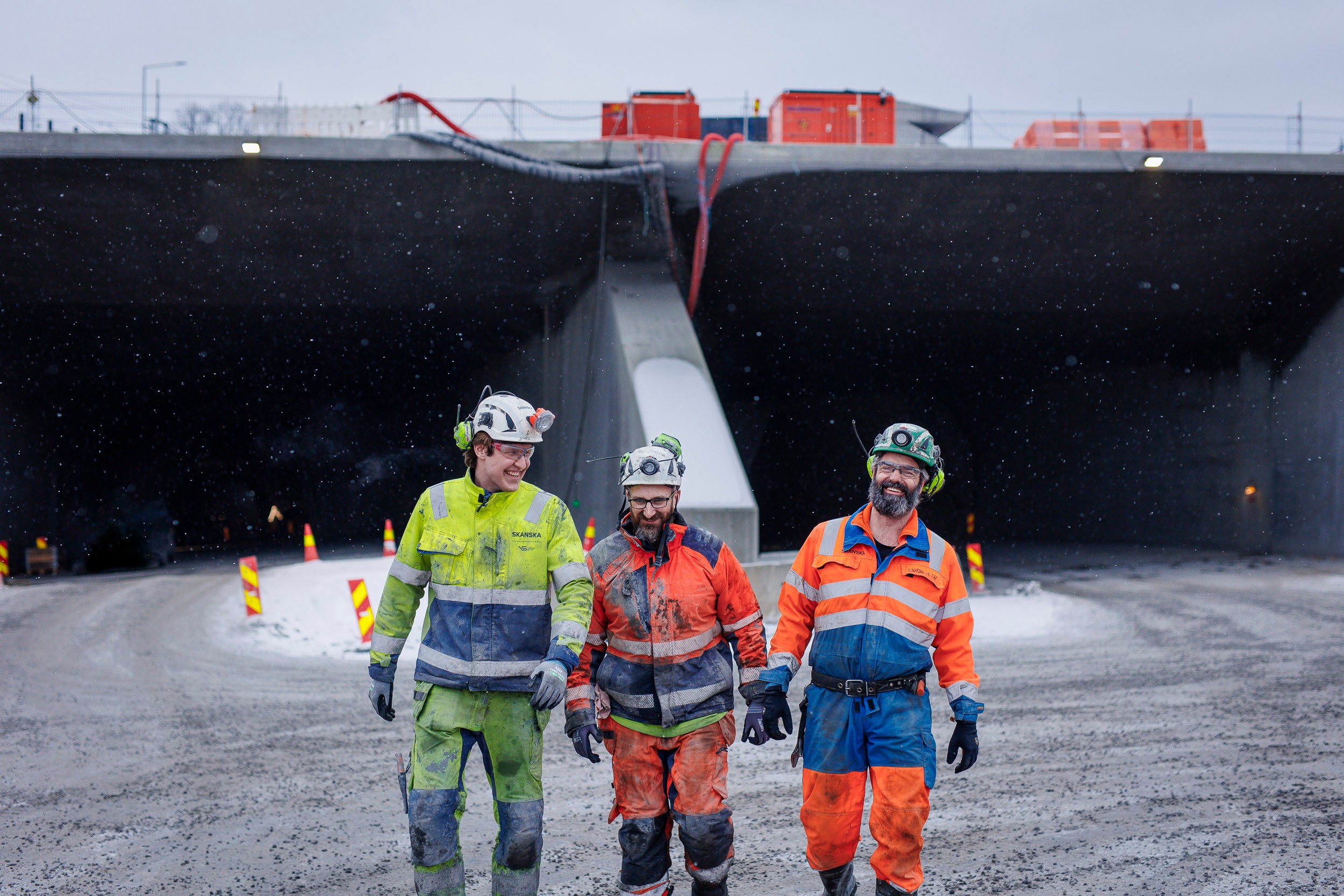 Construction Workers at Tunnel Site in Winter Conditions Tre byggearbeidere i verneutstyr går nær en betongkonstruksjon med maskineri bak.