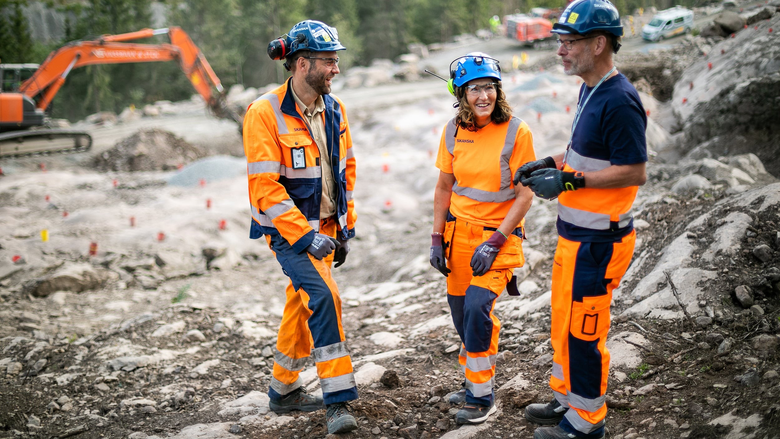 Construction workers in safety gear at an active site Tre bygningsarbeidere i verneutstyr som diskuterer på en steinete utendørs arbeidsplass.