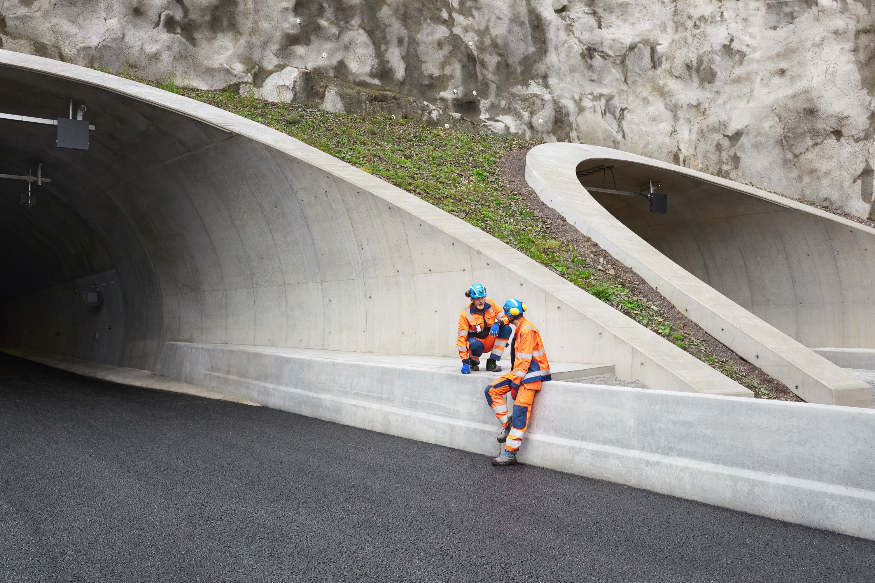 Tunnelarbeidere i sikkerhetsutstyr som hviler To bygningsarbeidere i oransje sikkerhetsutstyr hviler utenfor en moderne betongtunnelinngang.