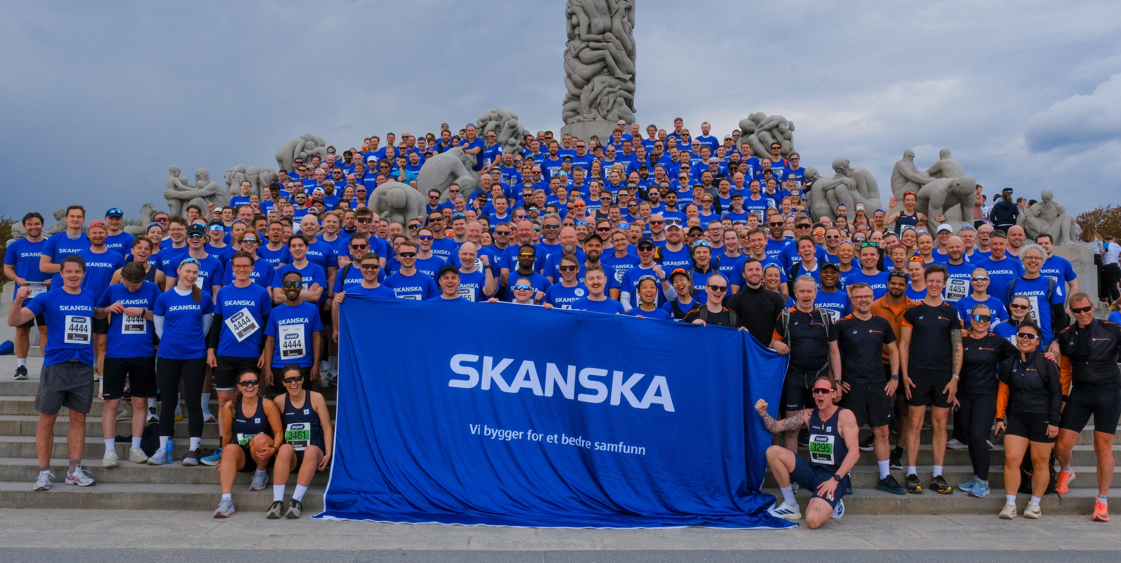 Skanska Team Gathering in Blue Shirts at Outdoor Monument Event Stor gruppe mennesker iført Skanska-skjorter samlet på trappene og holder et blått Skanska-banner.