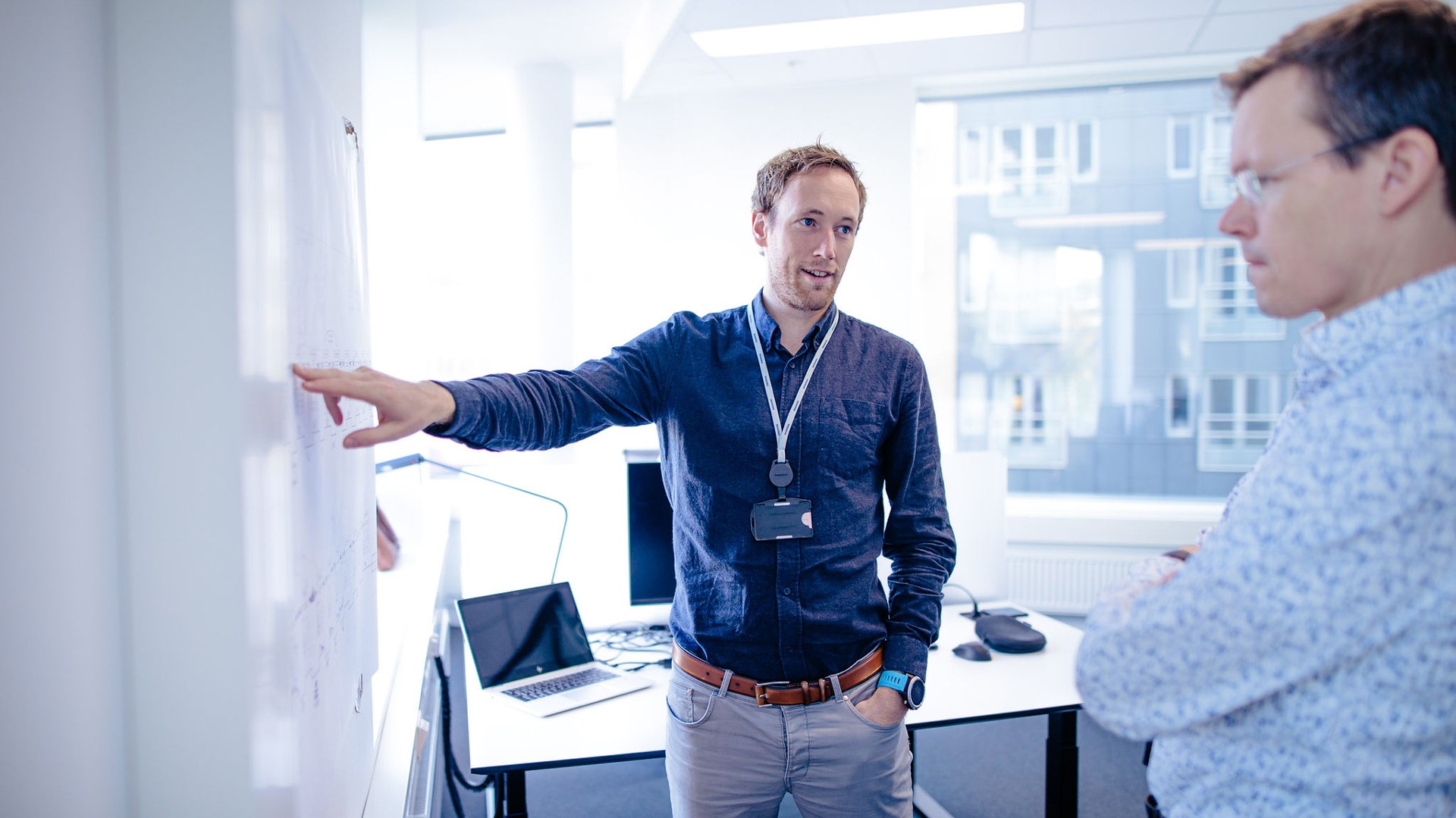Mann iført blå skjorte og smartklokke, stående med armene krysset i et lyst kontor.