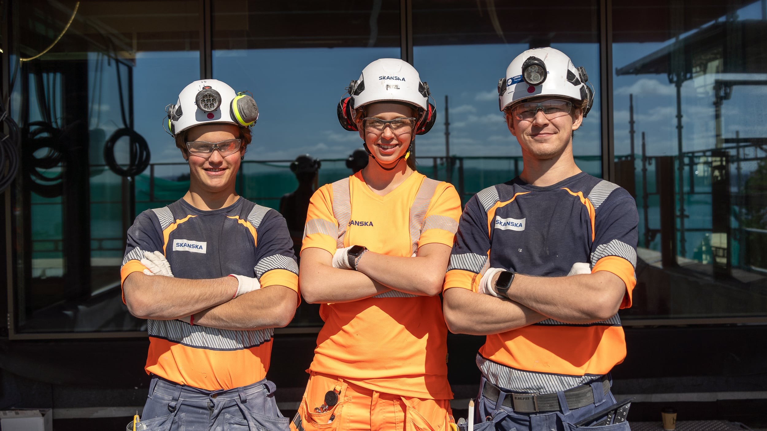 Construction Team in Safety Gear at Worksite Tre bygningsarbeidere i oransje sikkerhetsutstyr står med armene i kors på en byggeplass.