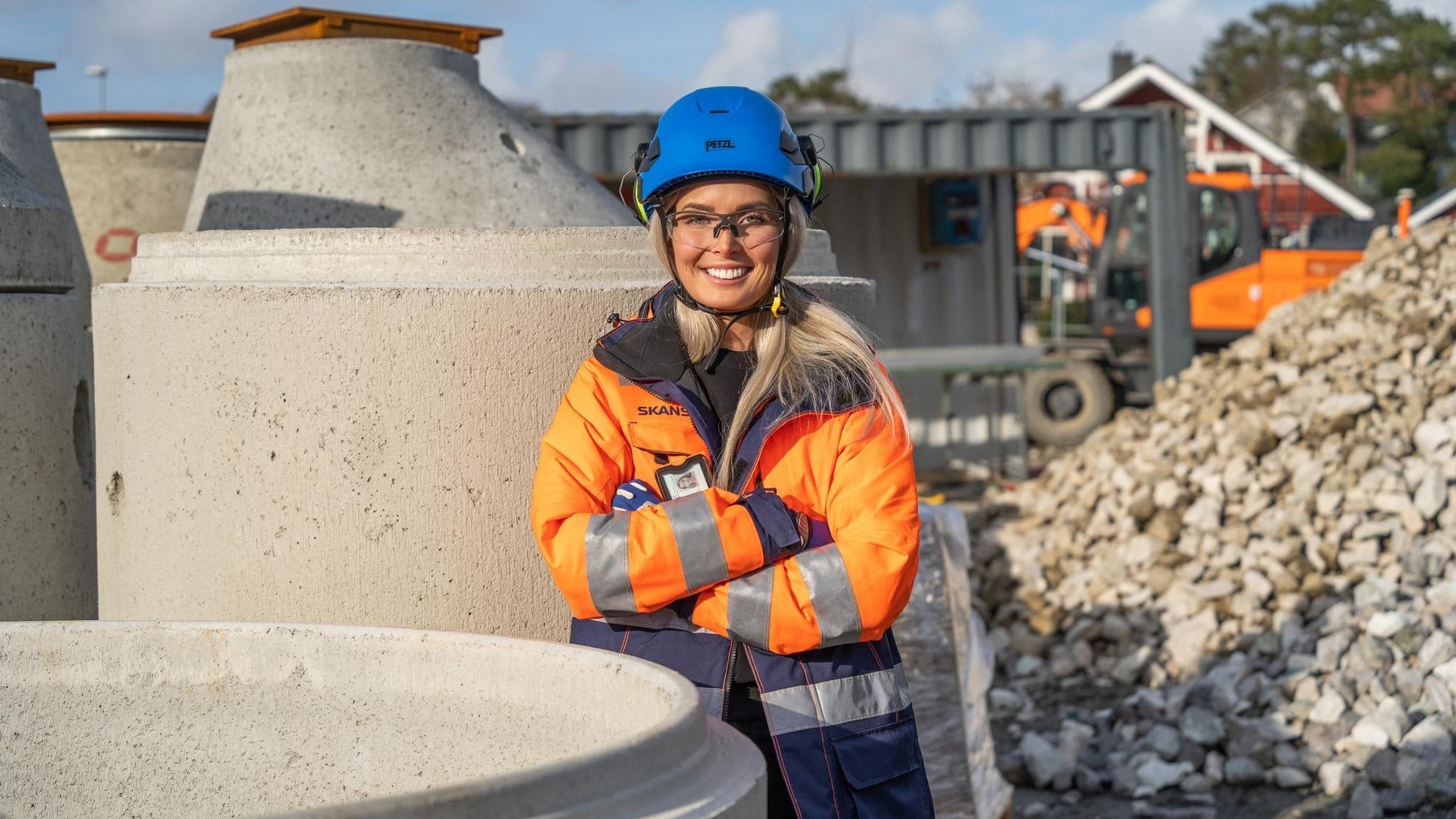 Female construction worker in safety gear at building site En bygningsarbeider i synlighetsutstyr står nær store betongstrukturer på en arbeidsplass.