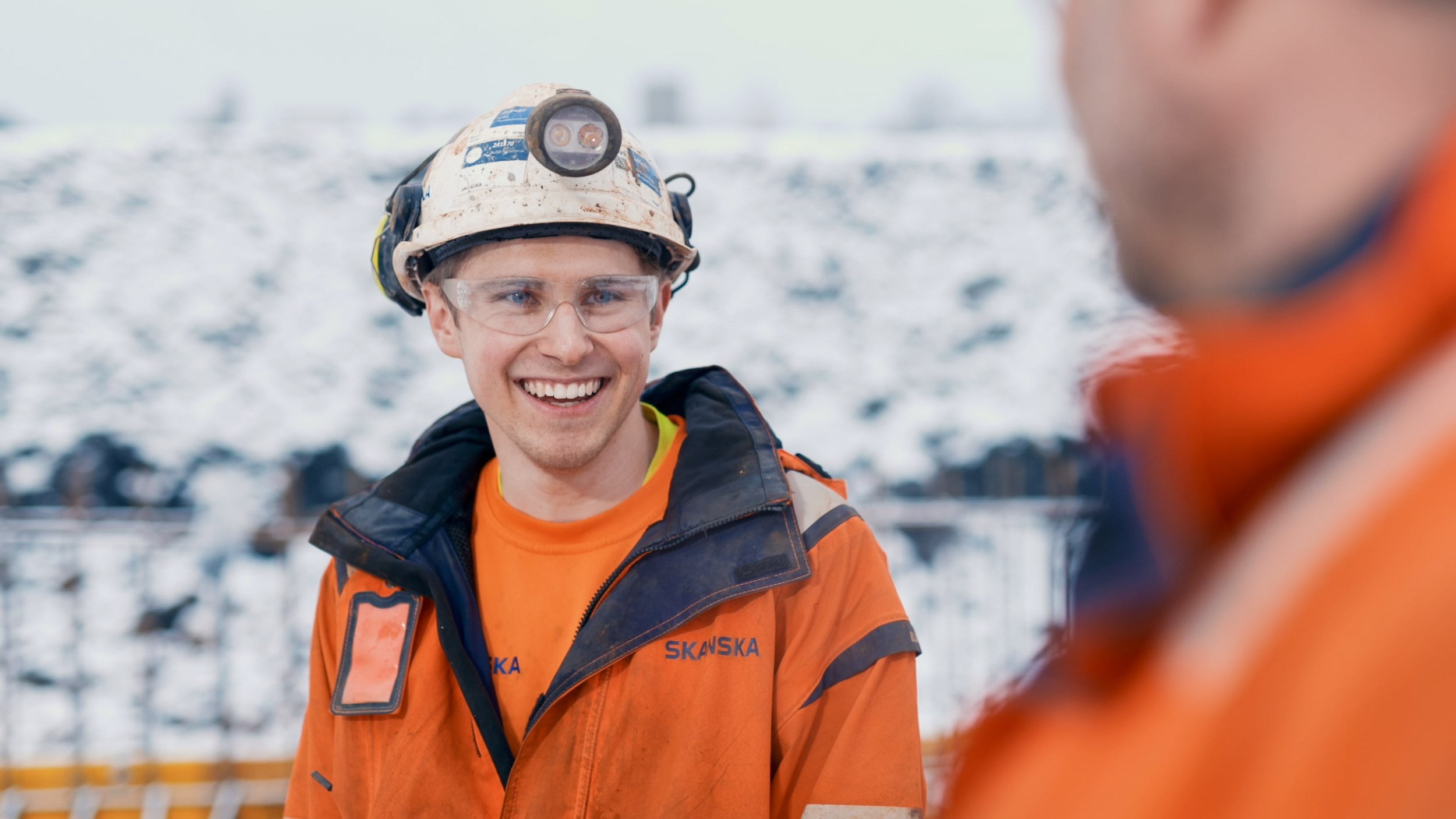 Construction worker in safety gear at snowy outdoor site Bygningsarbeider i oransje jakke og hjelm med lys på snødekt utendørs arbeidsplass.