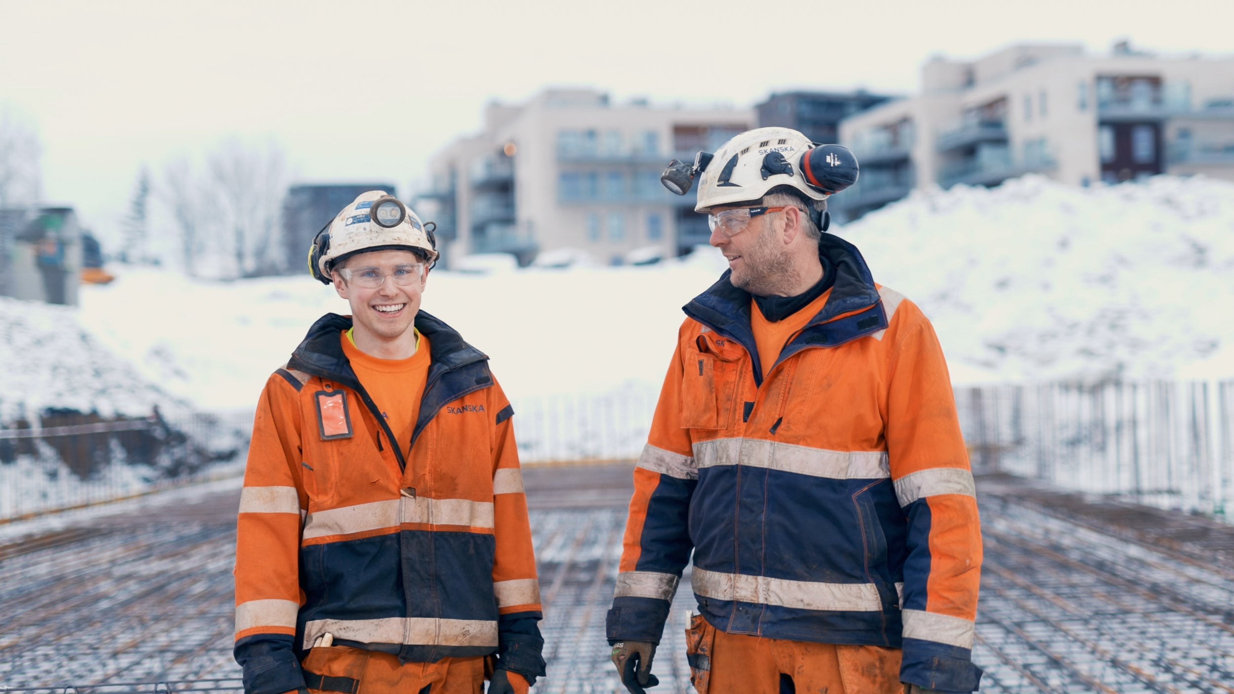 To bygningsarbeidere i oransje sikkerhetsutstyr og hjelmer står på en snødekt byggeplass.