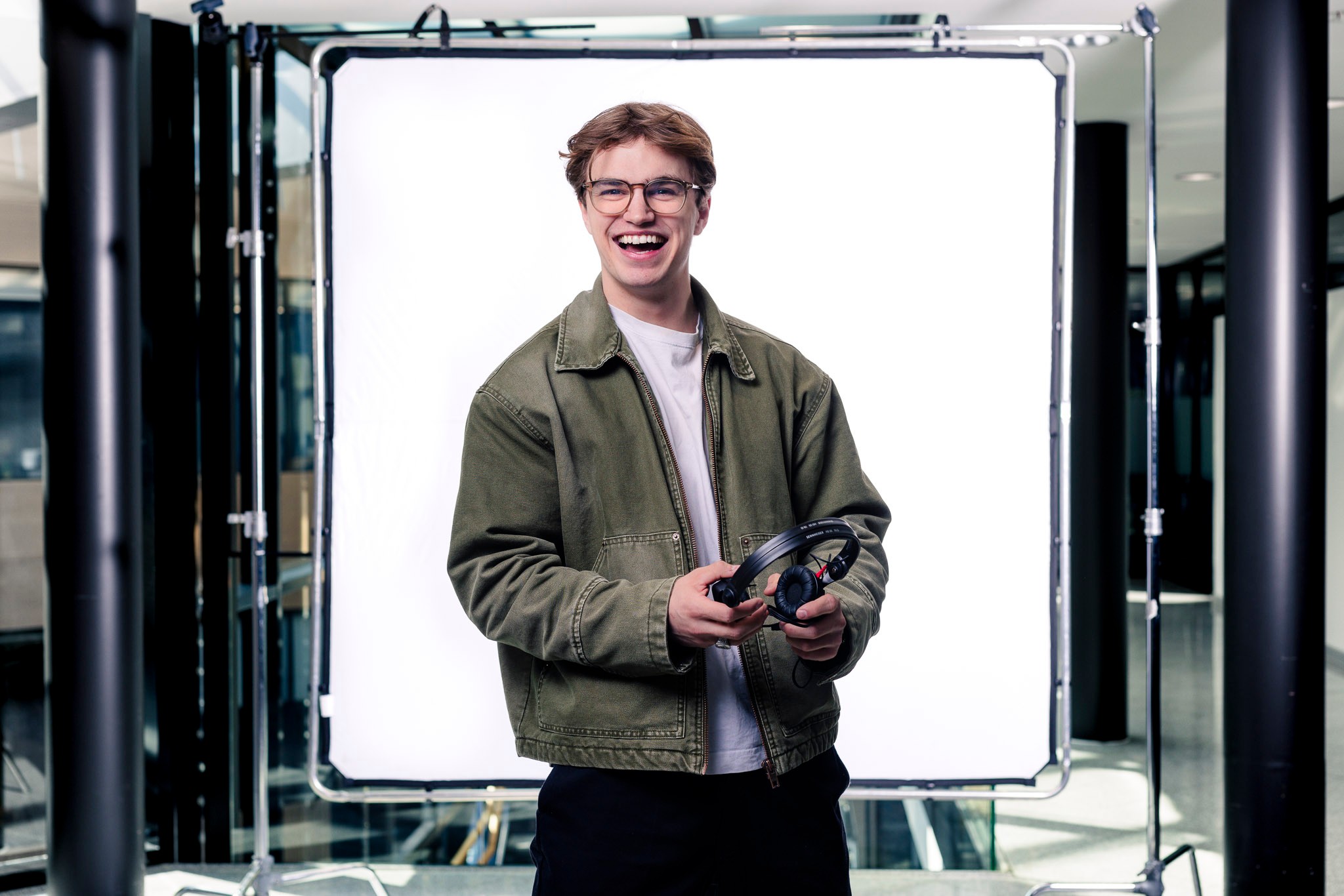 Man Holding Headphones Wearing Casual Jacket in Studio Setup Ung mann i grønn jakke som holder hodetelefoner i moderne studio med lyst bakteppe.