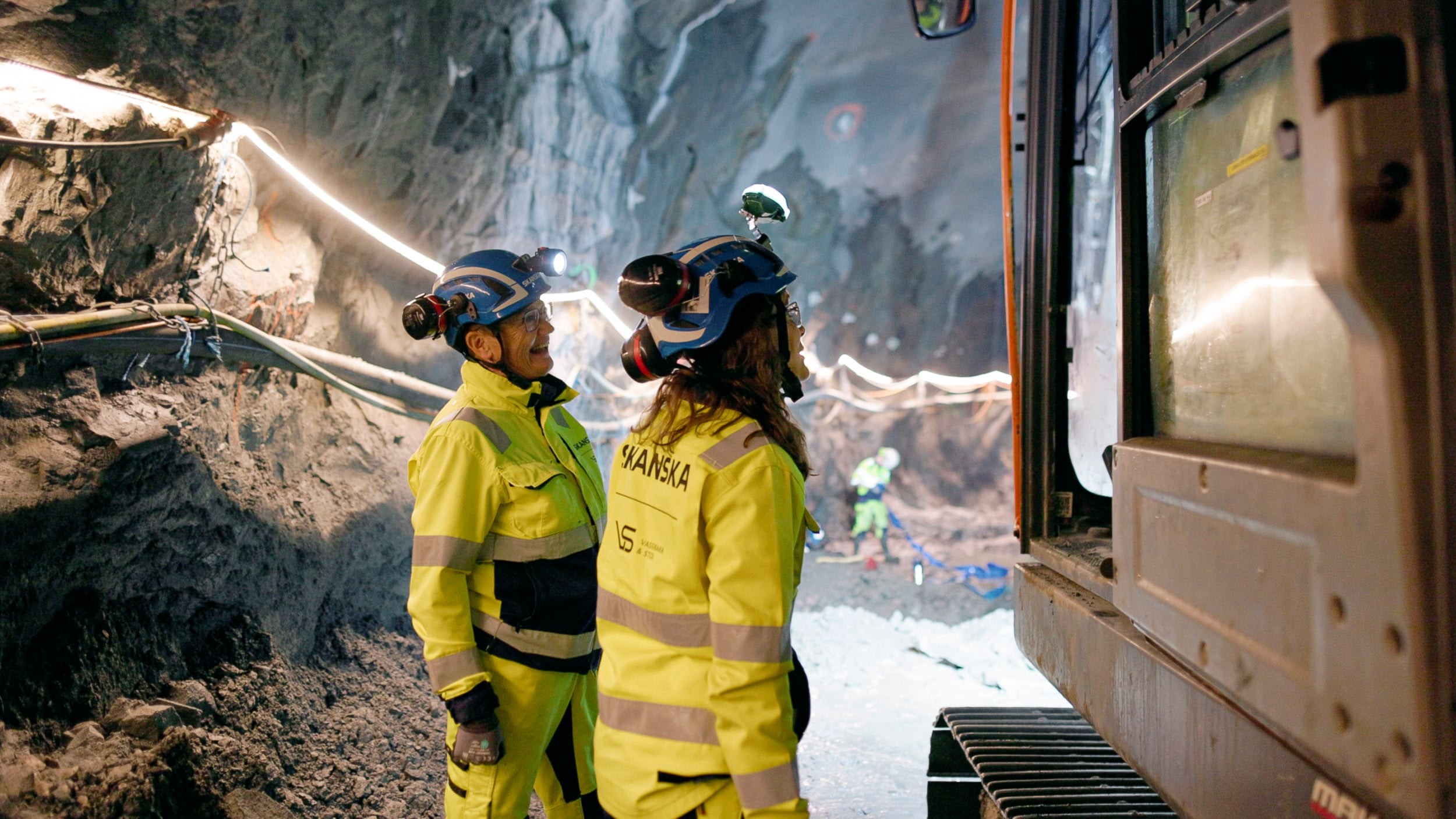 Underground Construction Workers Operating Heavy Machinery Bygningsarbeidere i gult sikkerhetsutstyr betjener maskiner inne i en steinete tunnel.