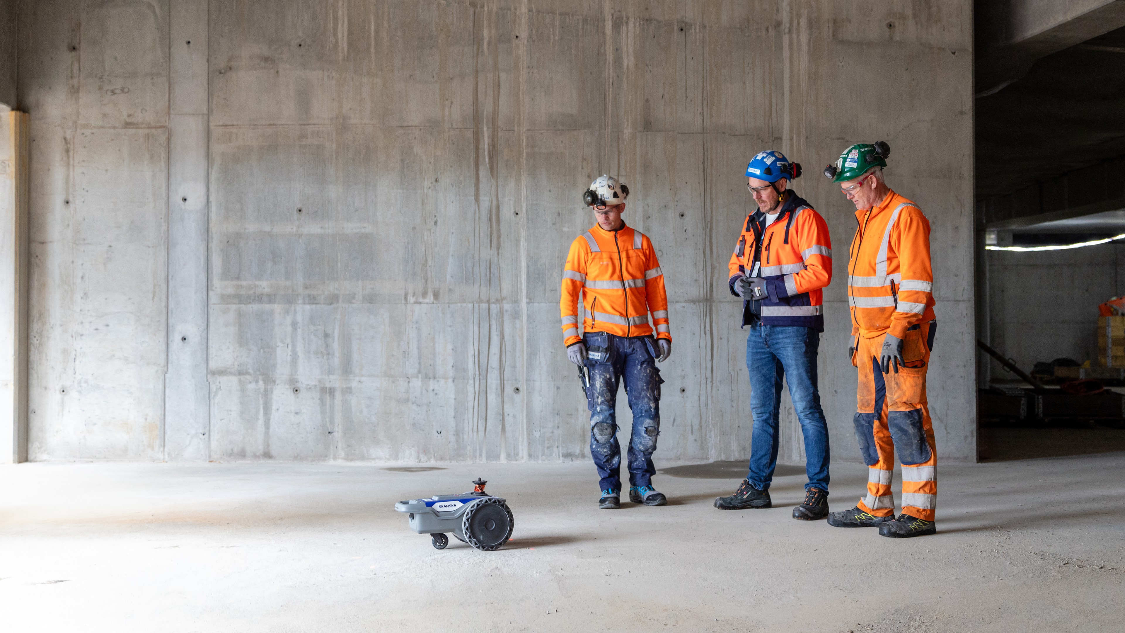 Bygningsarbeidere i sikkerhetsutstyr observerer en liten autonom robot på et betonggulv.