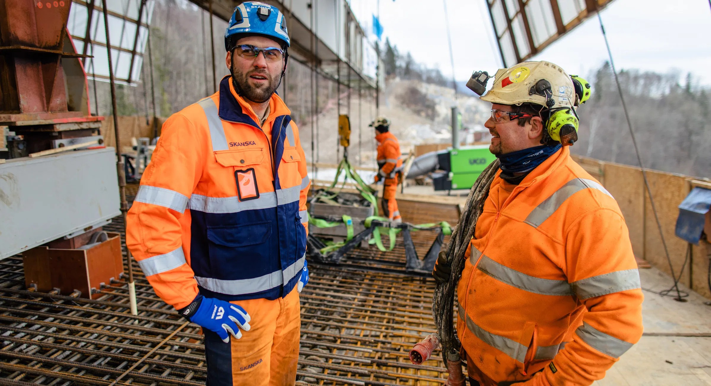 Bygningsarbeidere i oransje sikkerhetsutstyr på en byggeplass med stålramme.