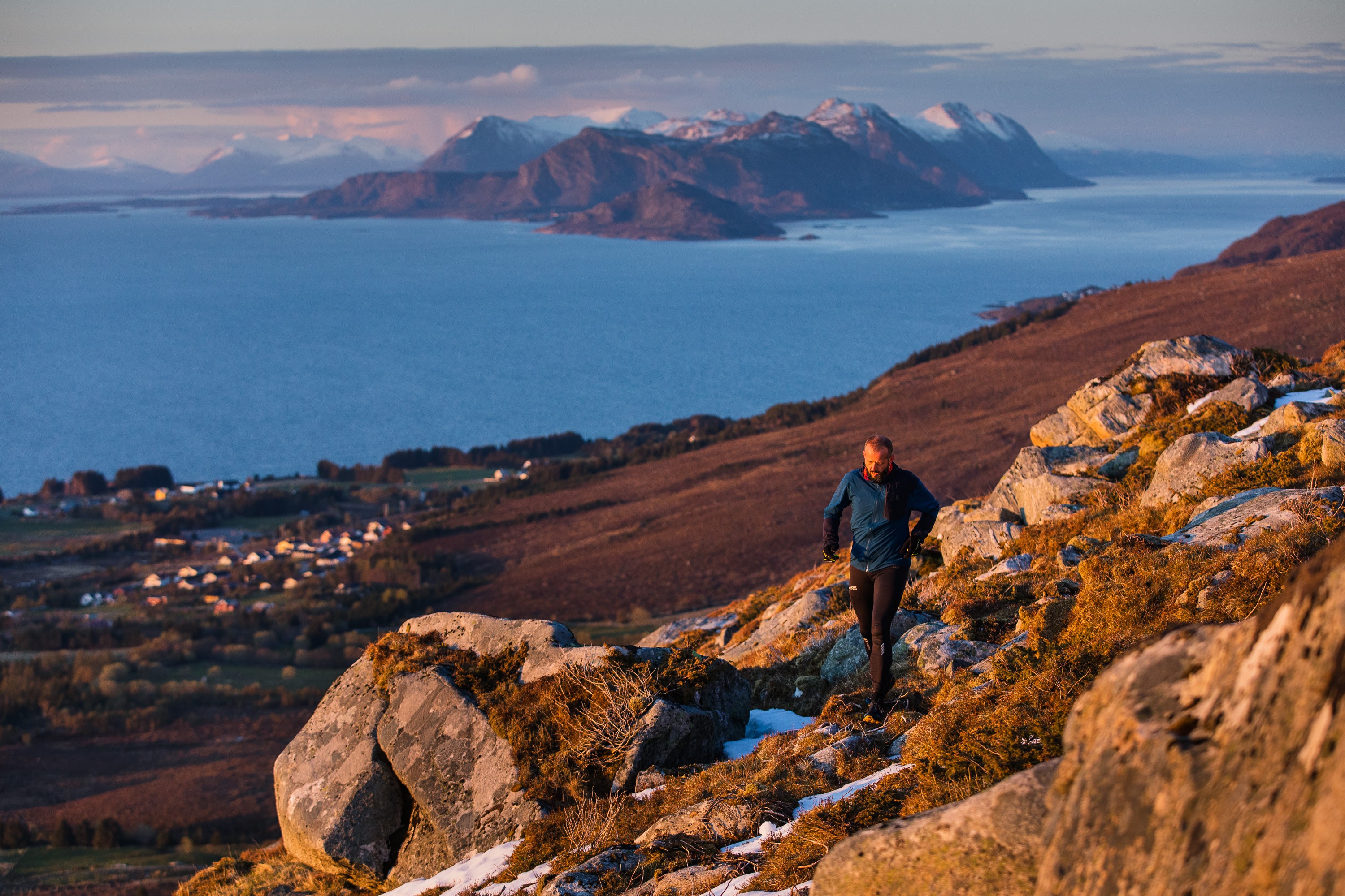 En person jogger på en steinete åsside med utsikt over et kystlandskap med fjell og en landsby nedenfor.