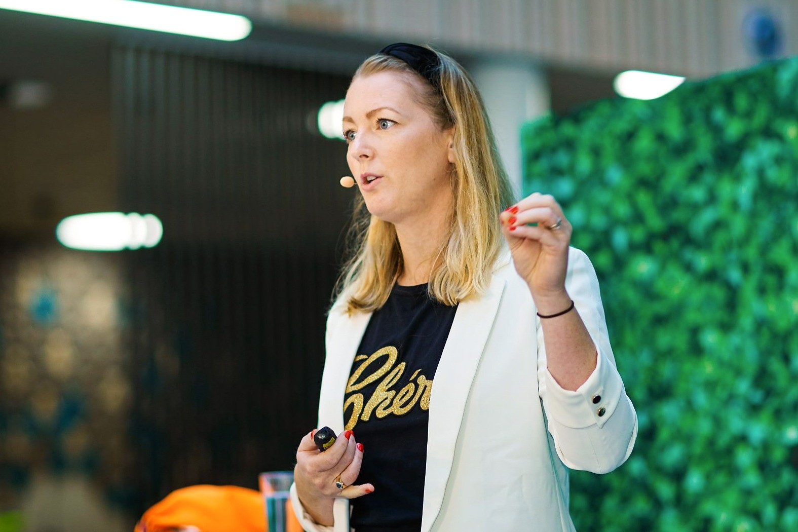 Kvinne som holder en presentasjon i stilig antrekk En kvinne i en hvit blazer gestikulerer under en presentasjon i en moderne, grønn-tematisert setting.