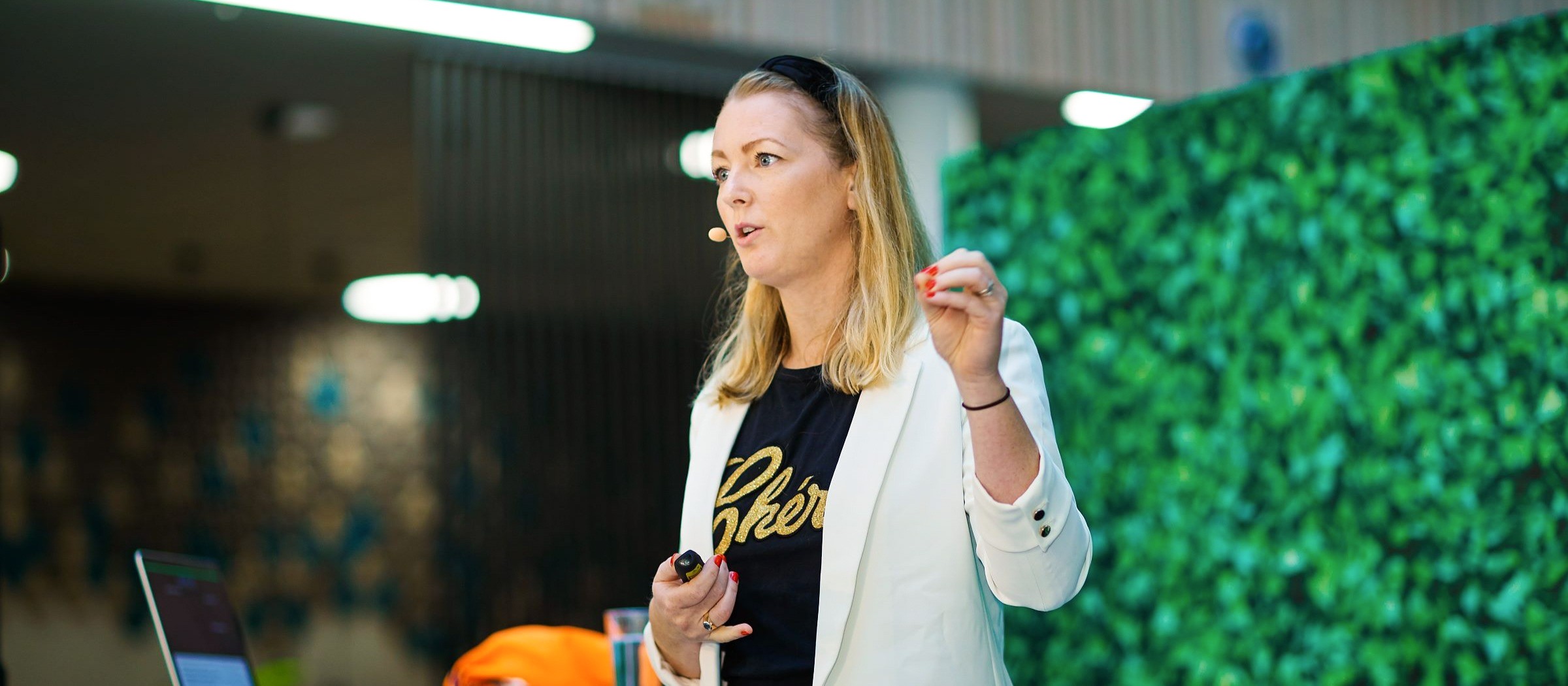 Profesjonell kvinne gir engasjerende forretningspresentasjon En kvinne i en hvit blazer holder en presentasjon i en moderne, grønn-tematisert setting.
