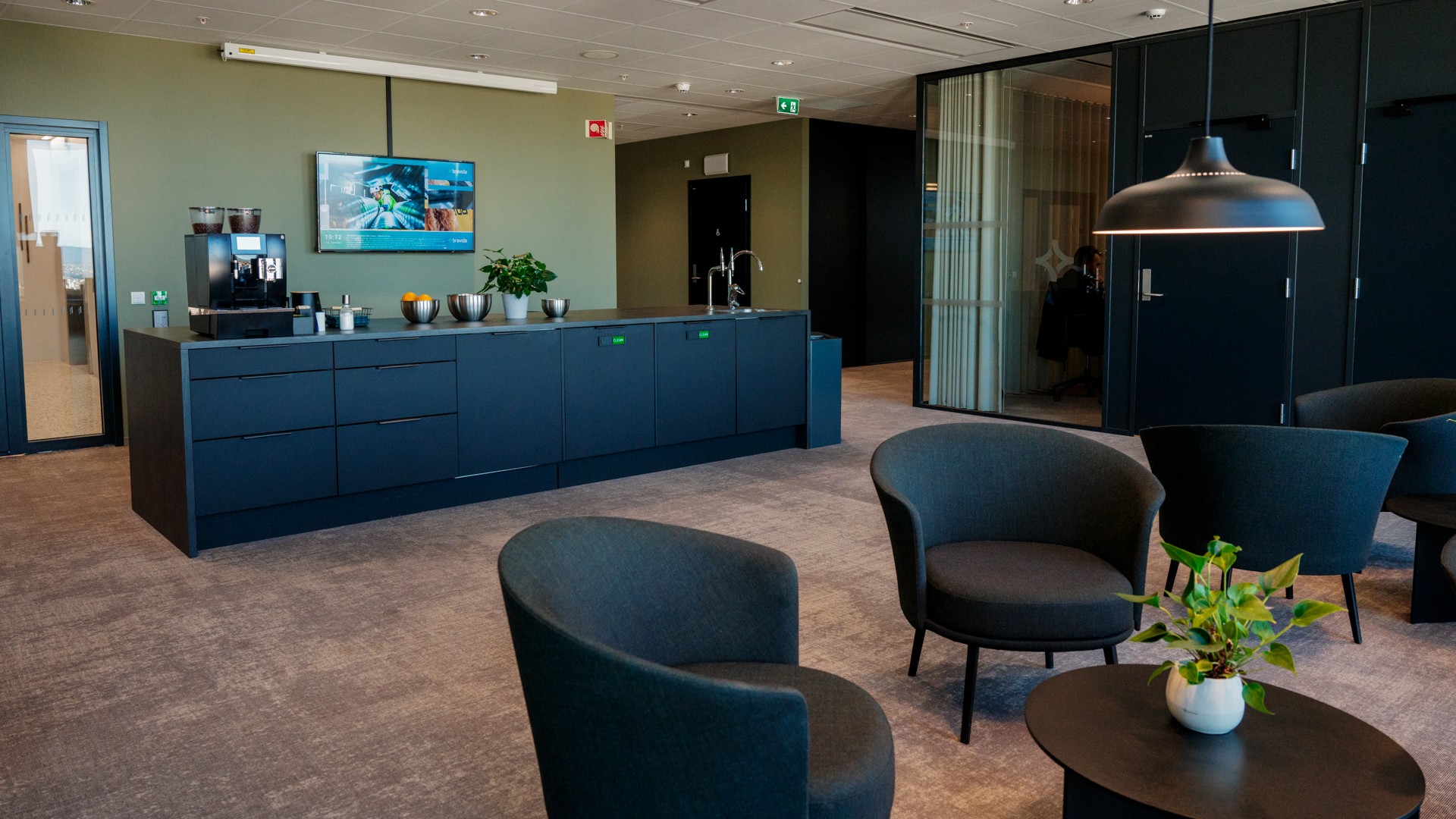 Moderne kontorlounge med stilige møbler, kaffestasjon og en stor veggmontert skjerm.