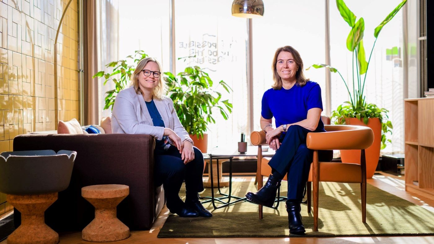 To personer sitter i et moderne, plantefylt kontorområde med stilige møbler og naturlig lys.