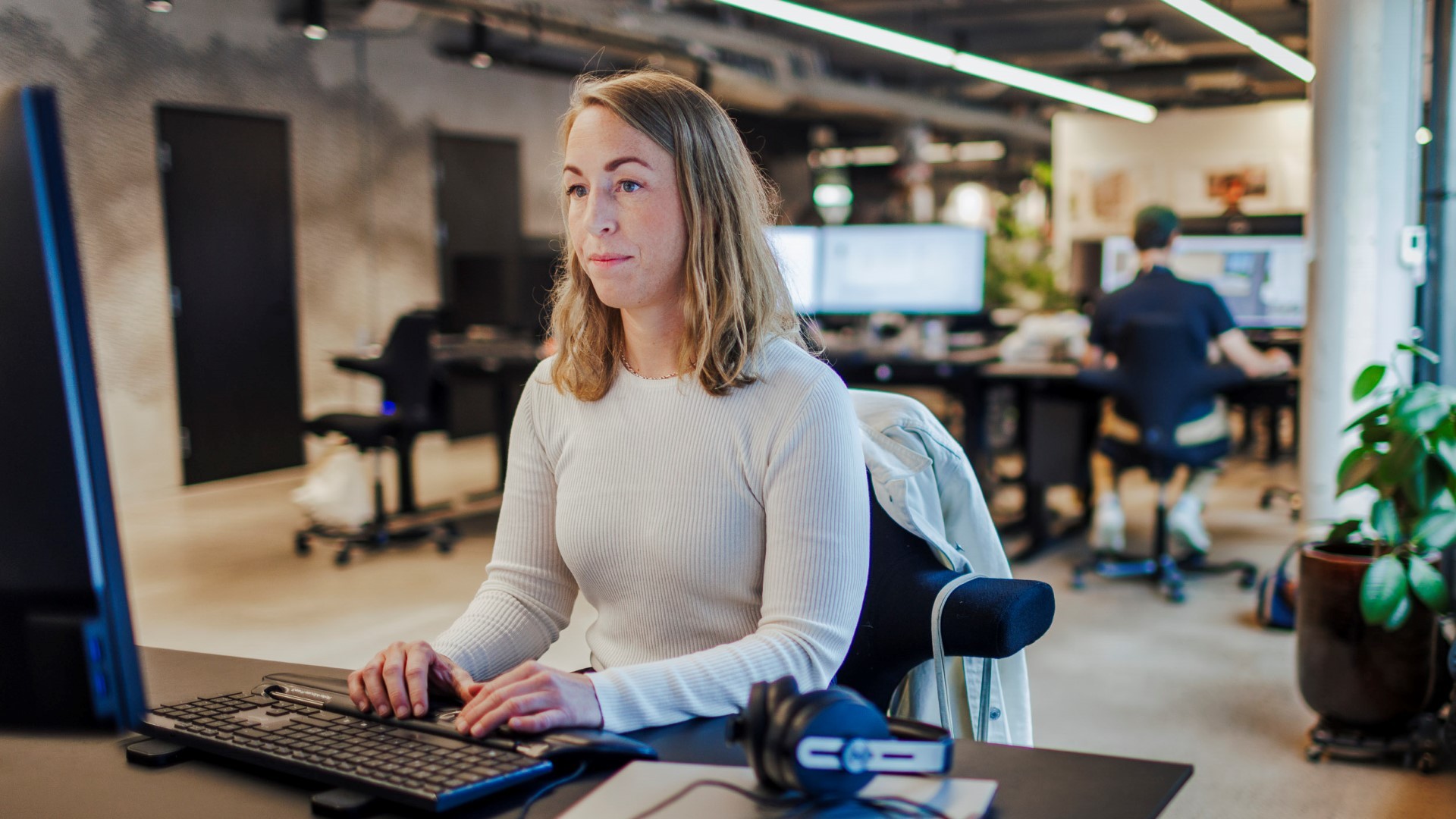 Moderne kontorarbeidsplass med fokusert profesjonell ved skrivebordet Kvinne som jobber ved en datamaskin i et moderne, åpent kontorlandskap med kolleger i bakgrunnen.