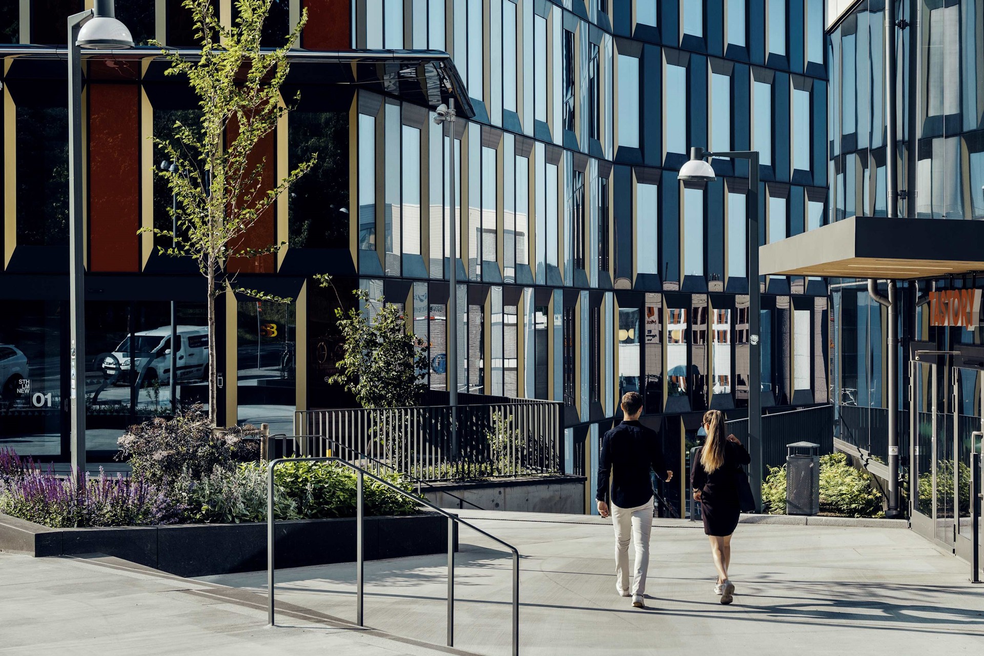 Moderne urban arkitektur med folk som går To personer går langs en moderne urban gate omgitt av glassbygninger og grøntområder.