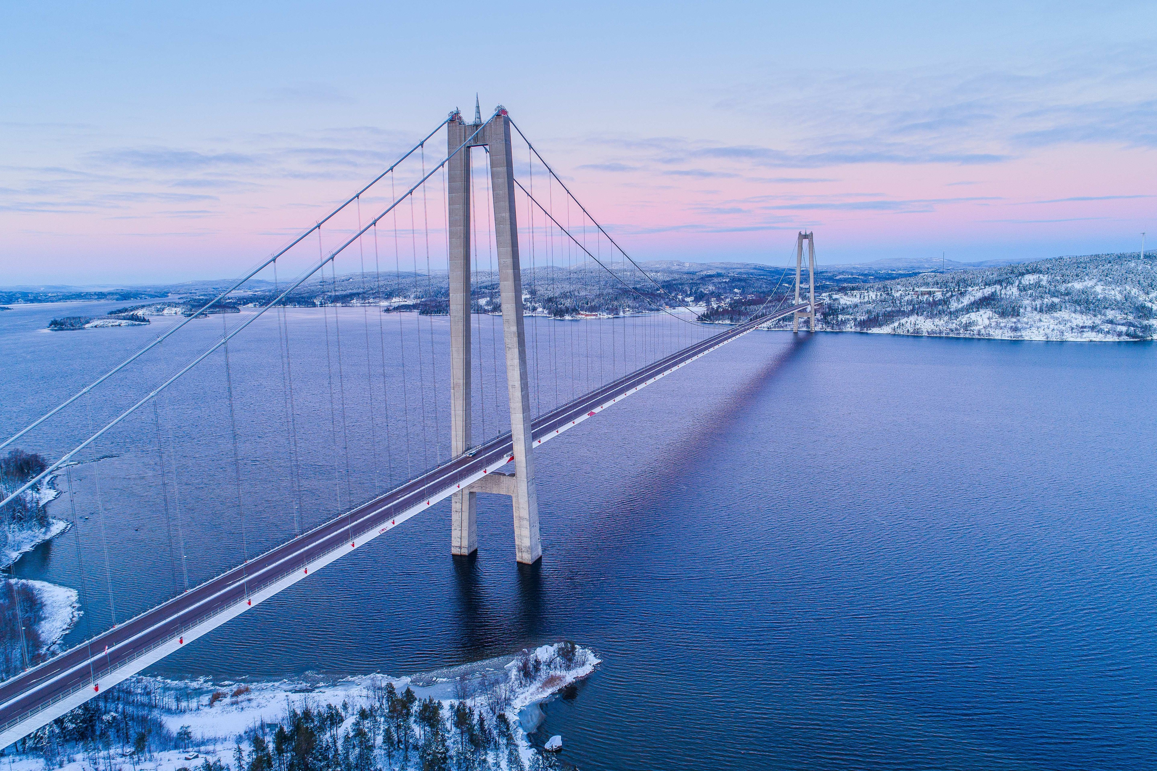 Högakustenbron Flygvy av en hängbro som sträcker sig över ett snöigt landskap över en lugn vattenyta vid skymning.