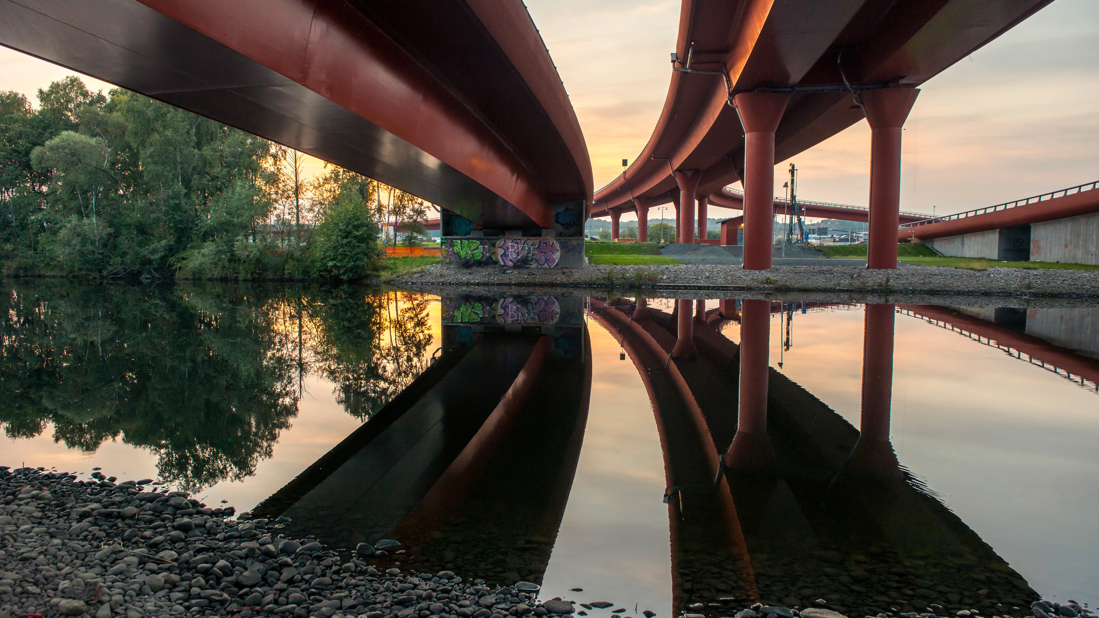 Upphöjda motorvägar reflekterade i en lugn flod, omgivna av träd vid solnedgången.