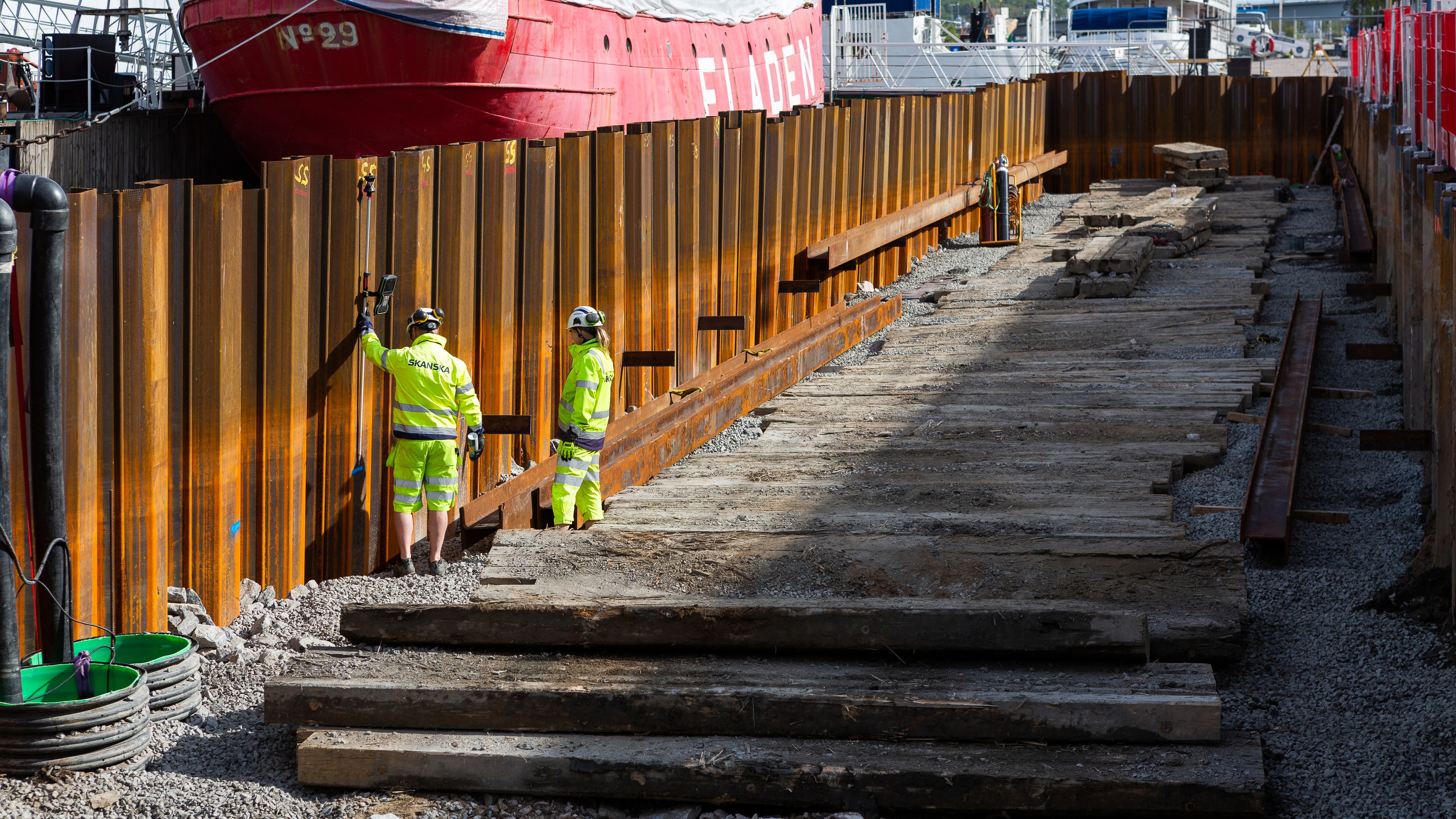 Byggnadsarbetare inspekterar stålspontvägg Två arbetare i skyddsutrustning inspekterar en byggarbetsplats med stålspont och träplankor.
