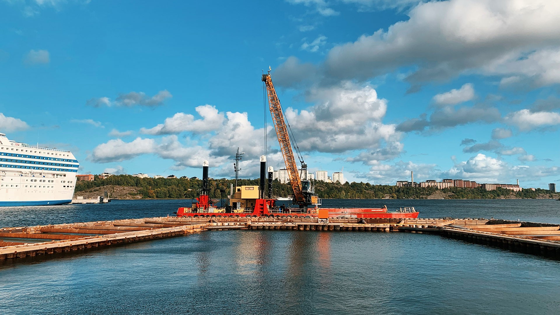 The Saltkajen construction site by the water with crane and ship in sight A construction crane on a platform by the water, with a cruise ship and cloudy sky in the background.