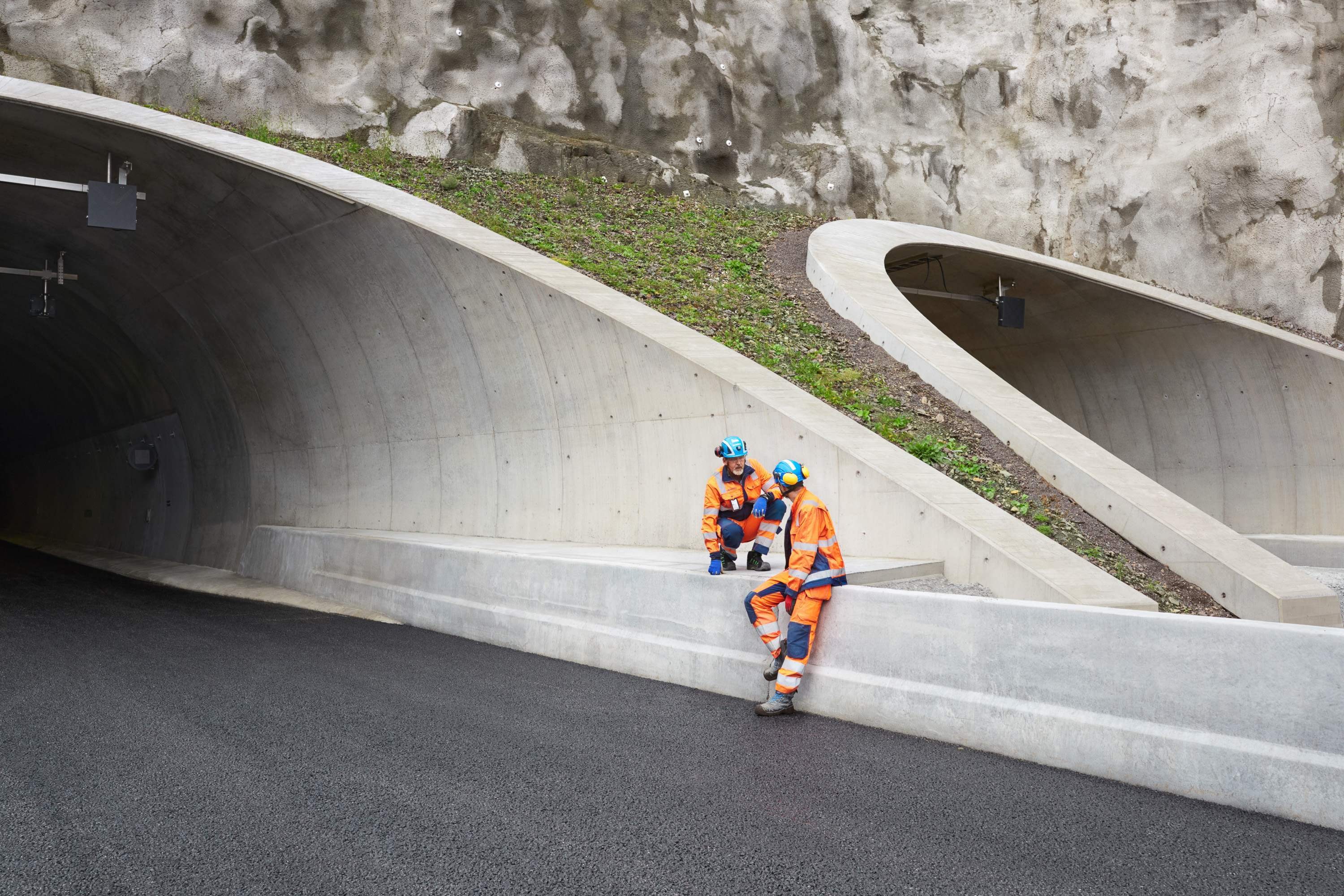 Tunnelbyggnadsarbetare i säkerhetsutrustning som vilar Två byggarbetare i orange säkerhetsutrustning sitter nära ingången till en betongtunnel.