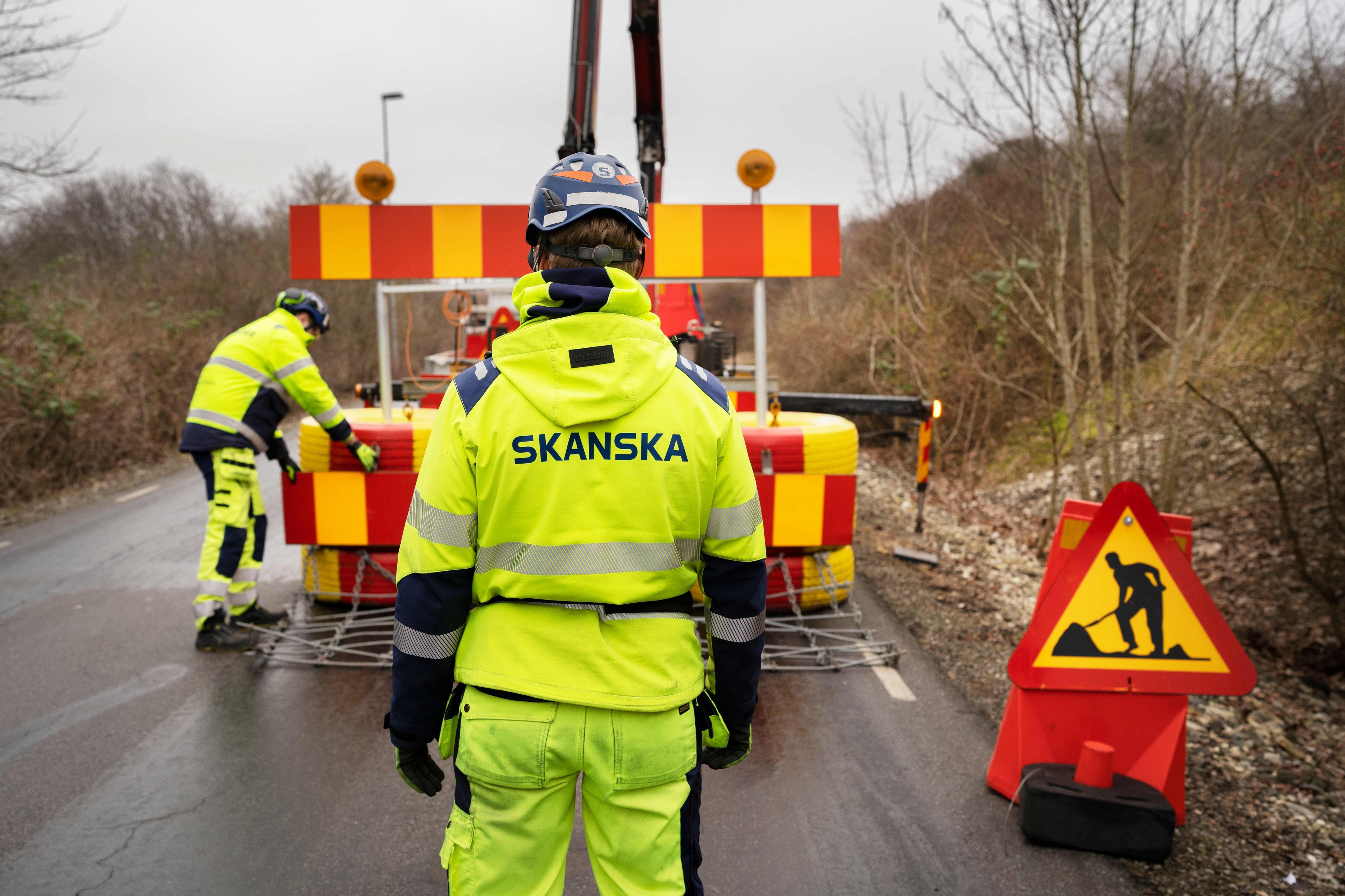 Vägarbetssäkerhet: Skanska-arbetare på platsen Byggnadsarbetare i högsynlighetskläder sätter upp vägbarriärer, märkta med en Skanska-logotyp.