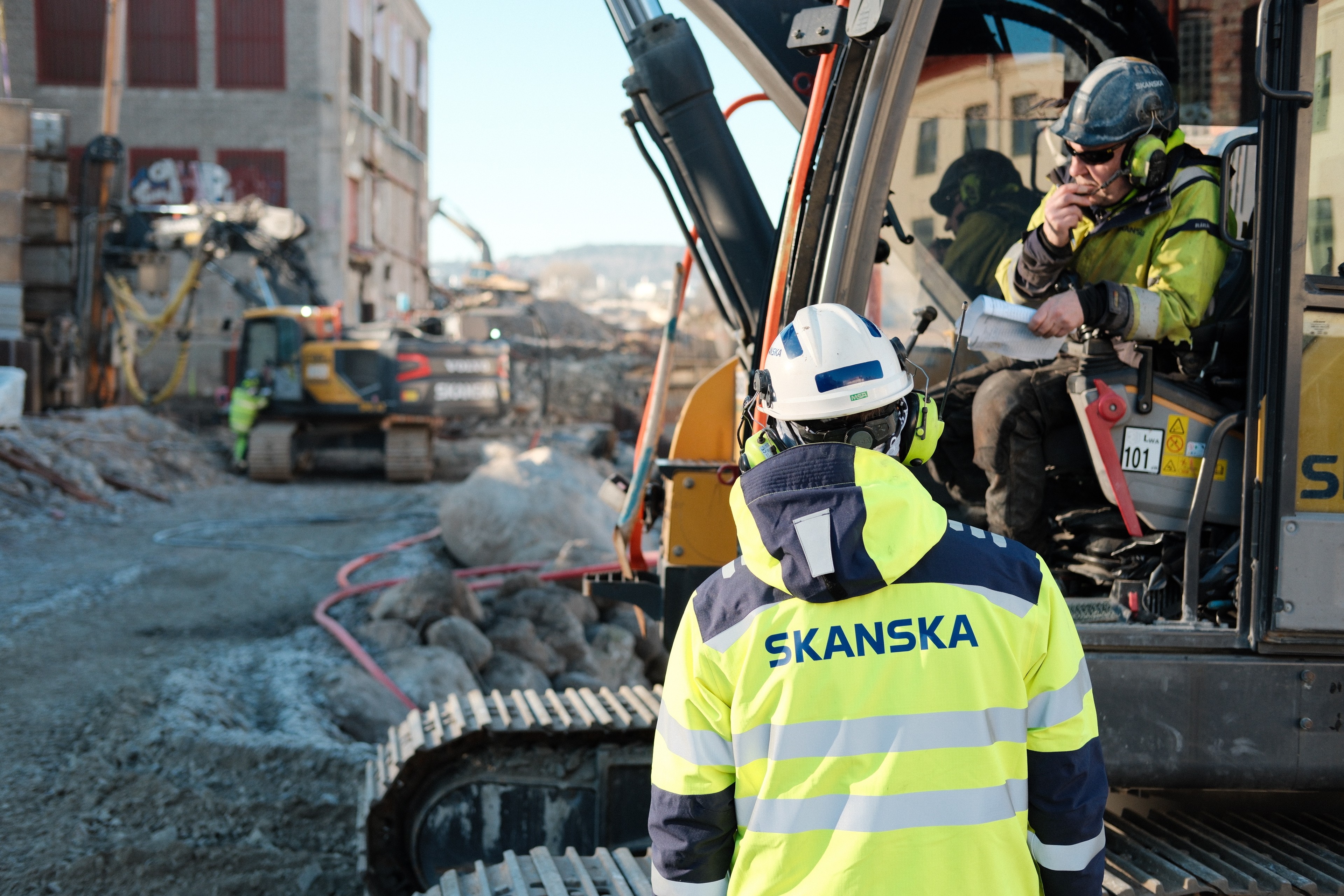 Vägarbetslag lägger asfalt med maskiner Byggnadsarbetare i Skanska-kläder på en aktiv arbetsplats med maskiner i bakgrunden.