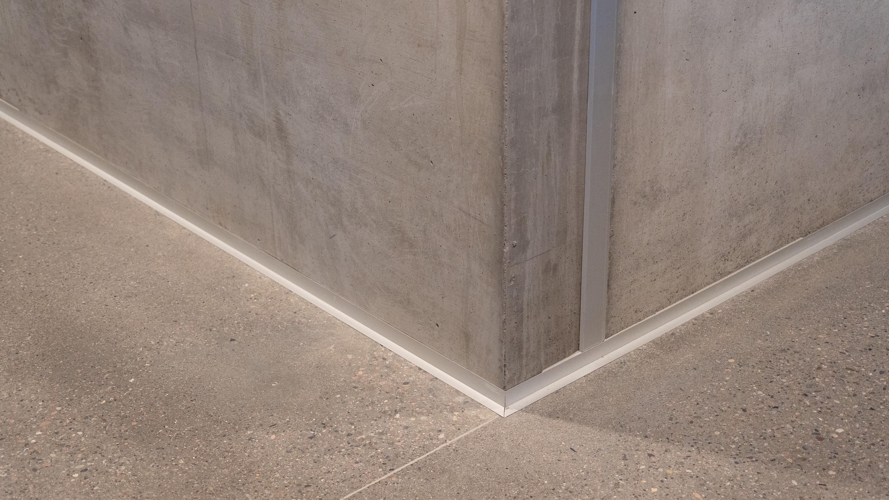 Modern concrete wall corner with clean white baseboard and polished stone floor.
