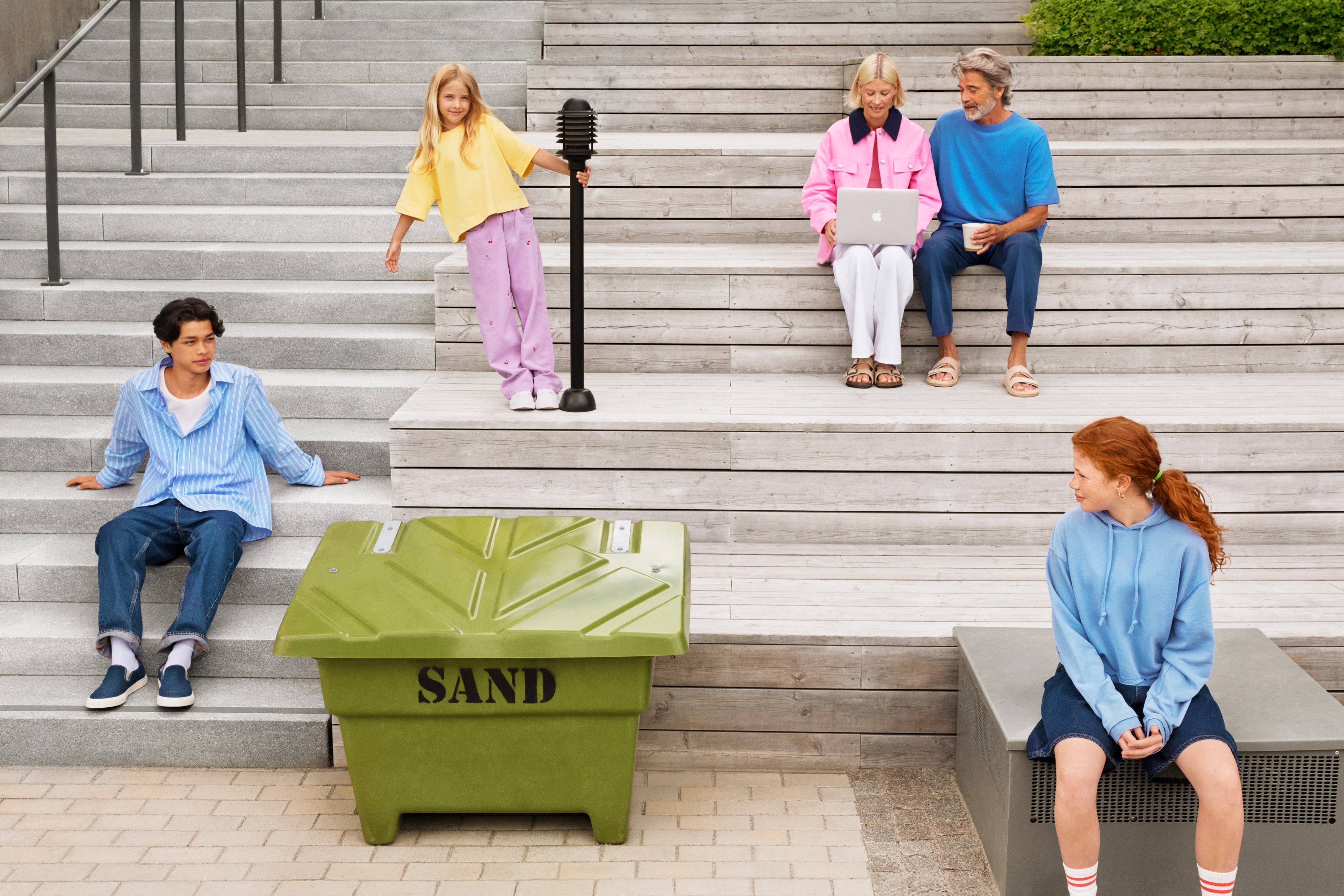 Modern utomhus urban livsstil med mångsidig grupp av människor En grupp människor slappnar av på utomhustrappor nära en grön sandlåda, en håller en laptop.