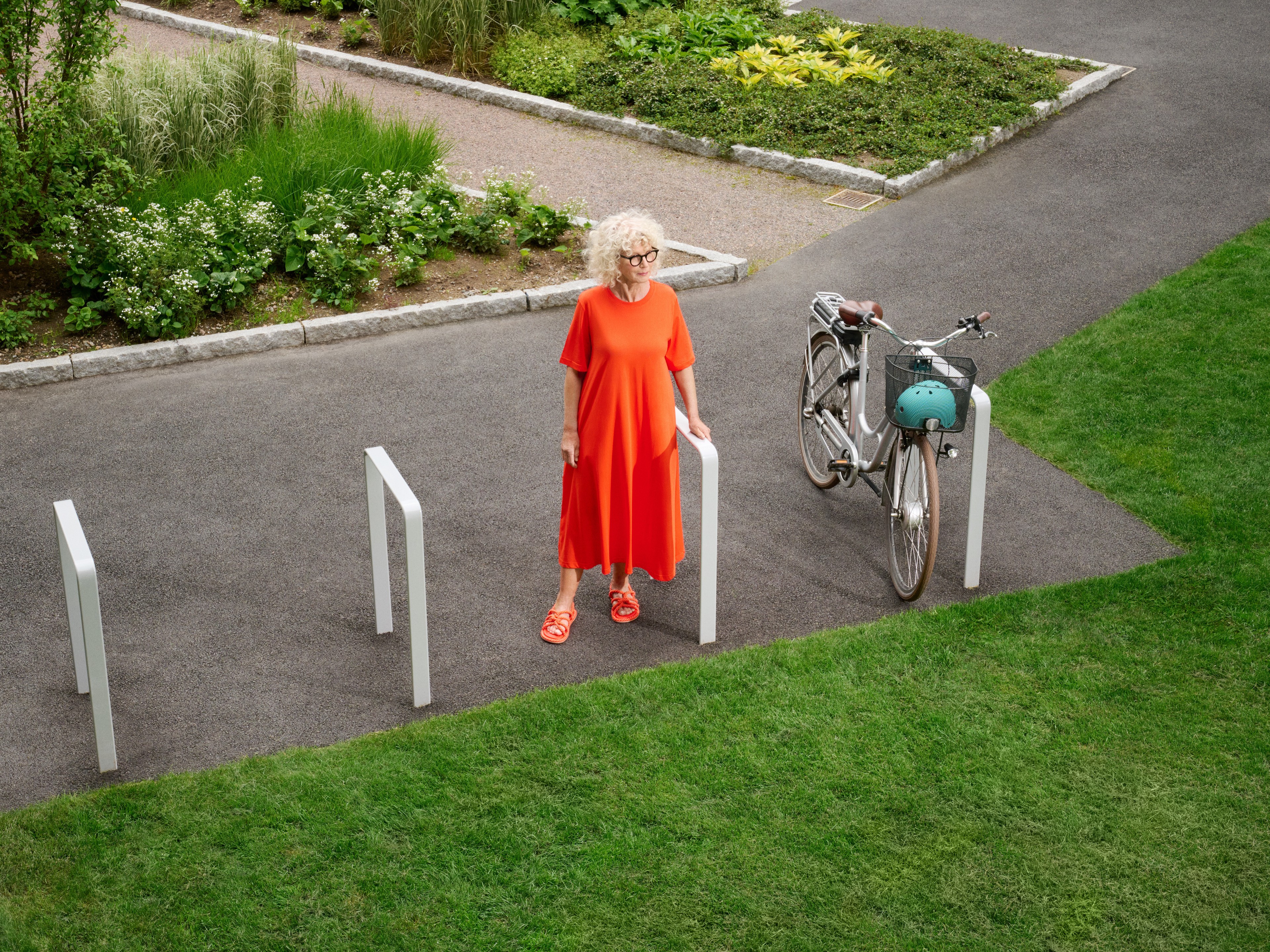 Kvinna i röd klänning som står nära en parkerad cykel på en asfalterad stig i en grön park.