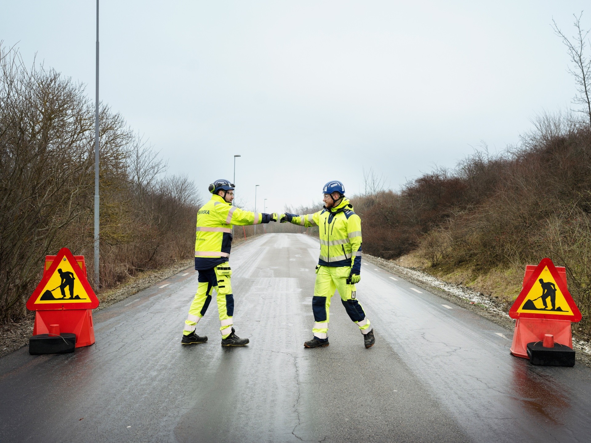 Two construction workers in high-visibility gear fist bump on a road between work signs.