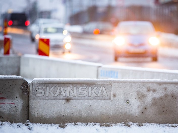 Snötäckt betongbarriär med "SKANSKA" branding på en trafikerad väg med suddiga bilstrålkastare.