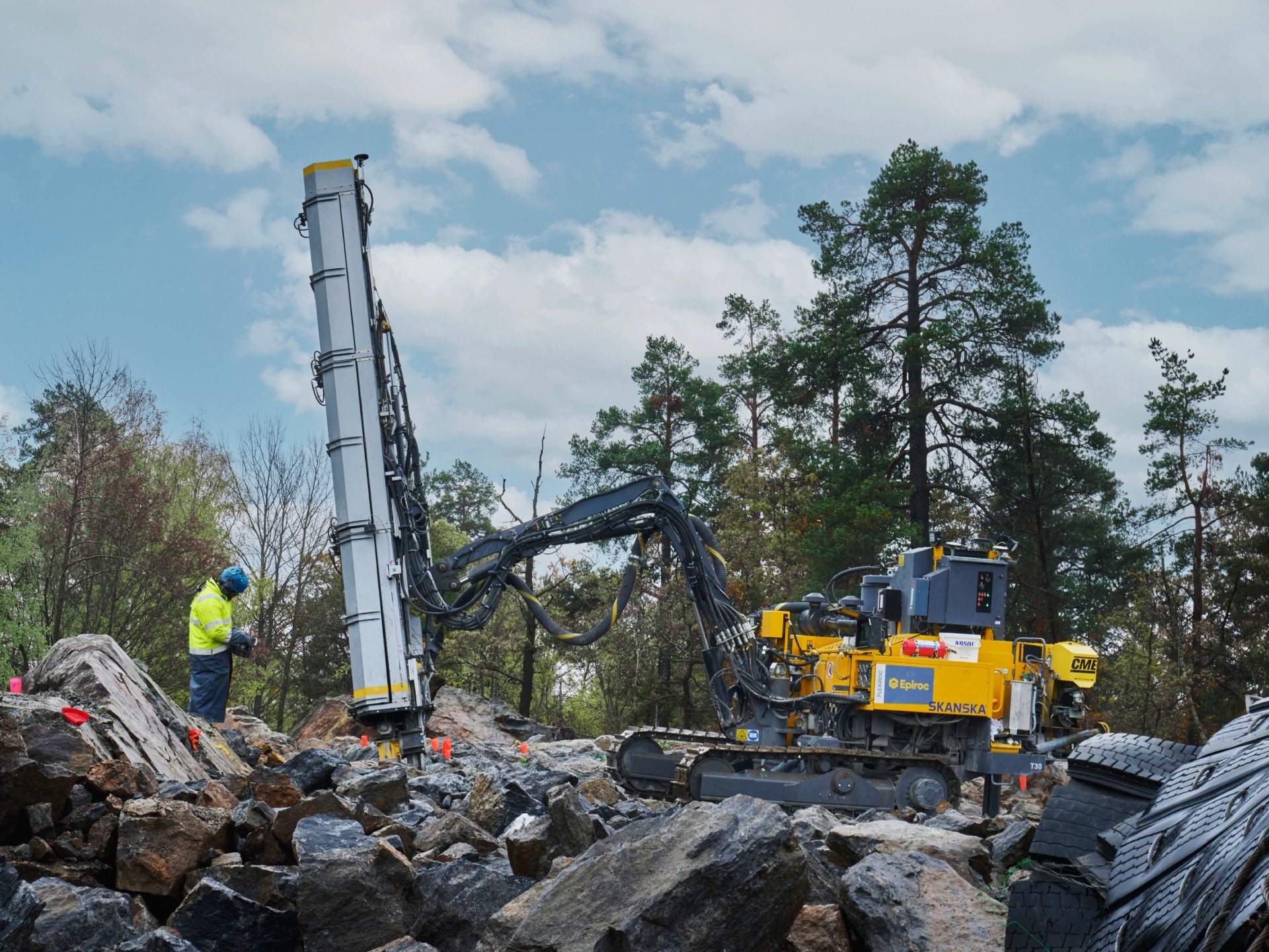 A construction worker operates a large drilling machine on a rocky terrain under a cloudy sky.