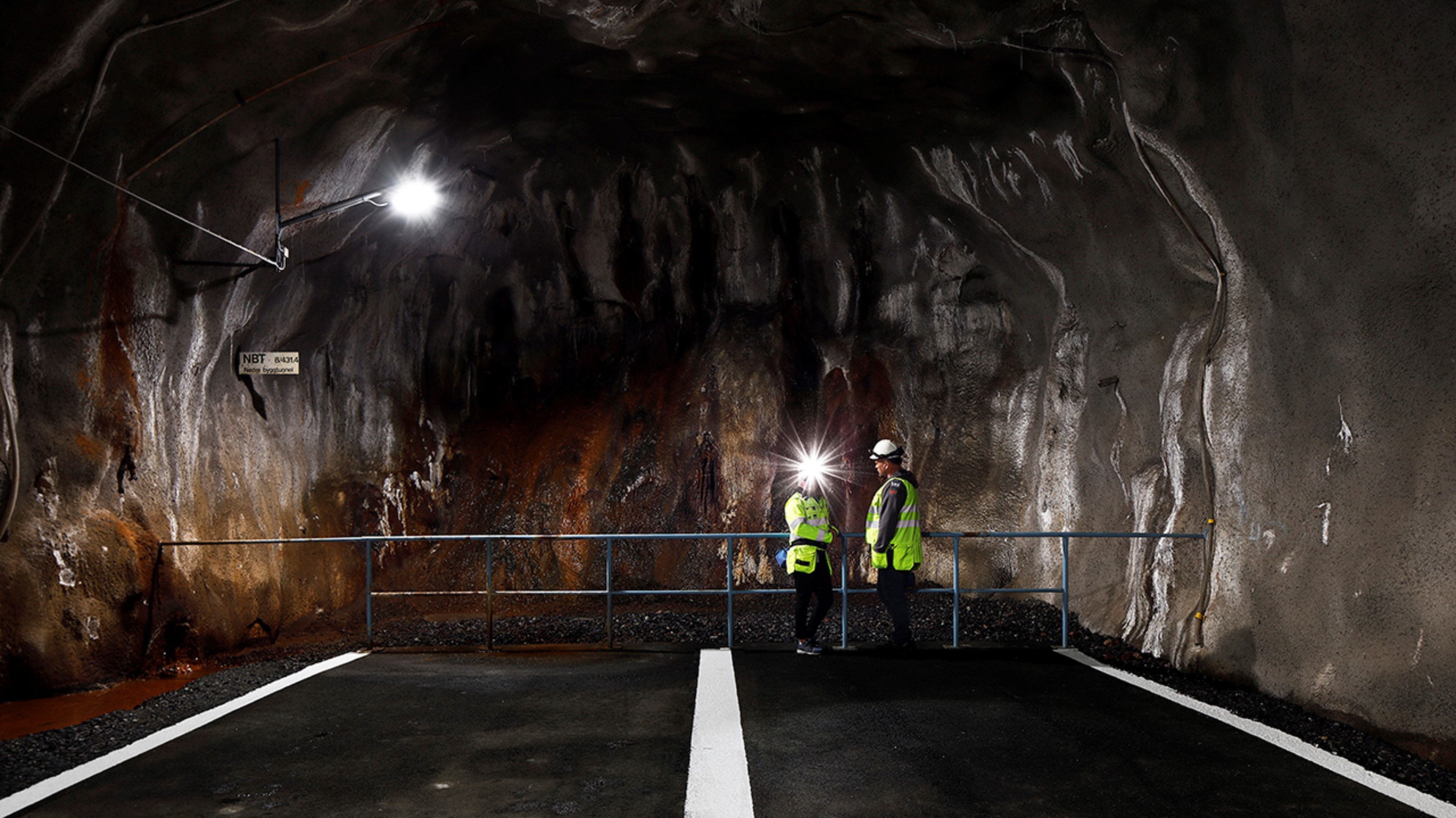 Två arbetare i säkerhetsutrustning står inne i en svagt upplyst underjordisk tunnel med klippväggar.