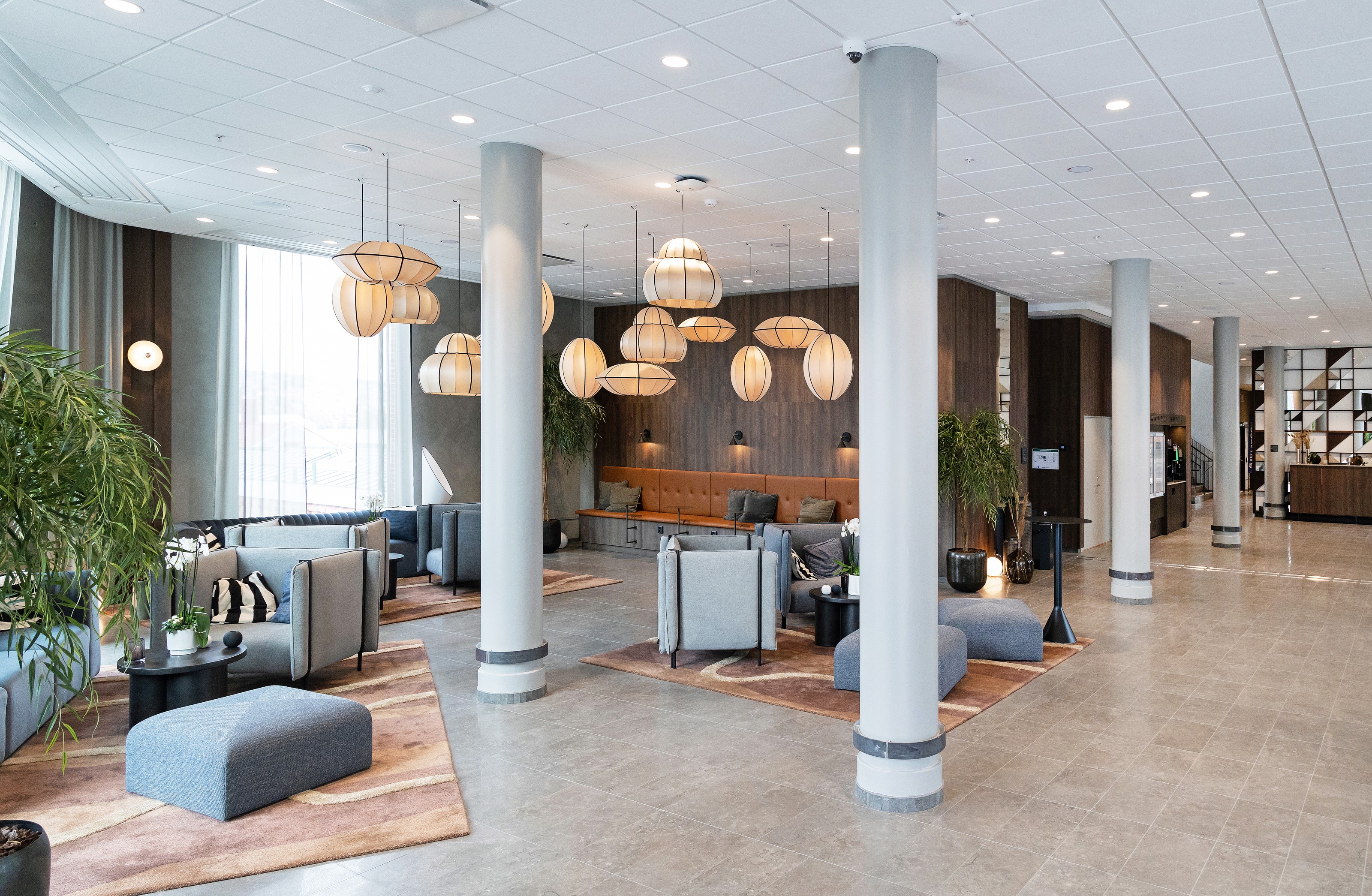 Modern kontorslounge med grå sittplatser, hängande lampor, växter och trädetaljer.