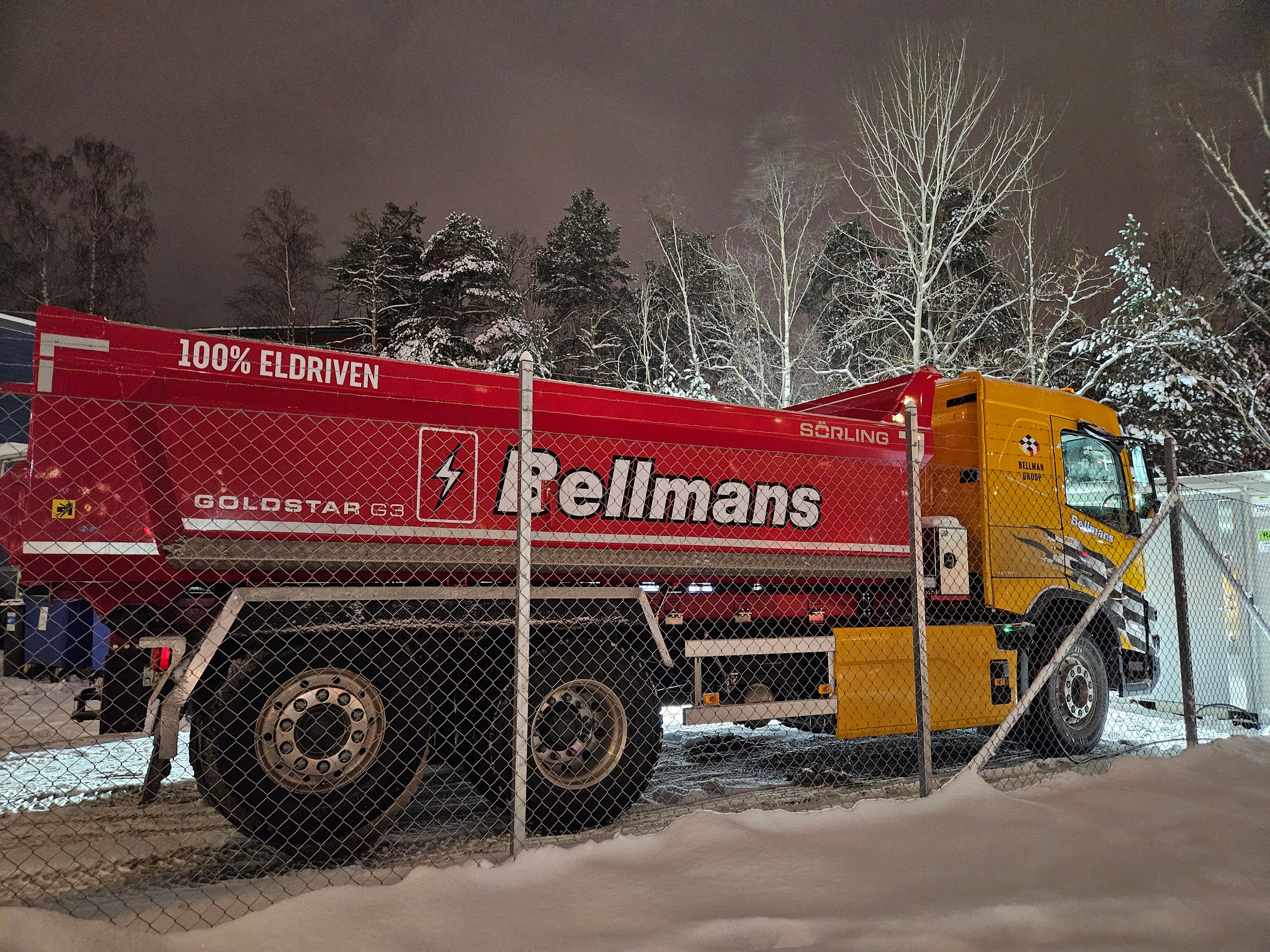 Röd Bellmans elektriska tipptruck parkerad bakom ett stängsel av kedjelänk i en snöig natt.
