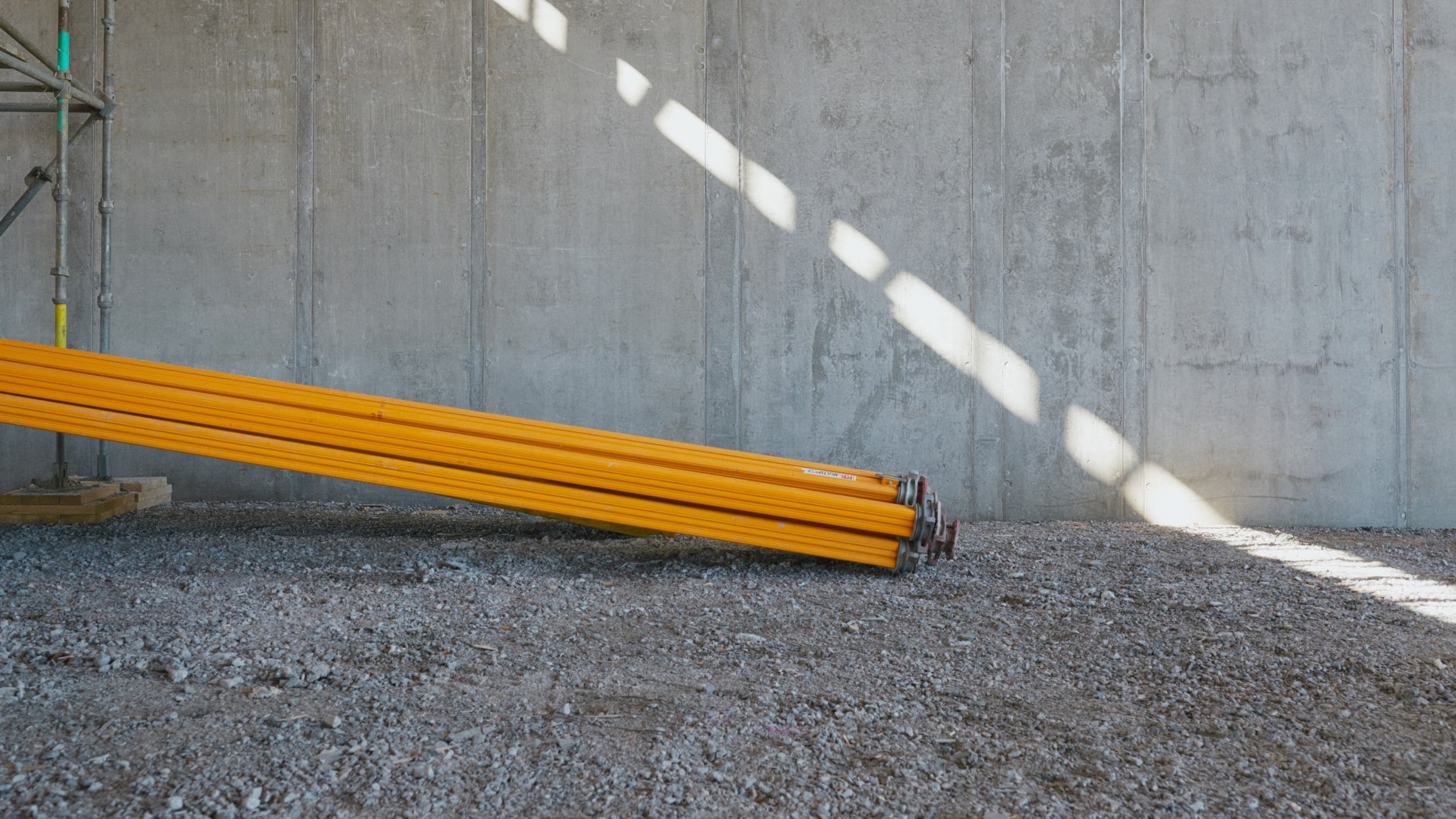 Construction Site with Yellow Steel Pipes in Sunlight Gula metallrör staplade på grus, lutade mot en betongvägg på en byggarbetsplats.