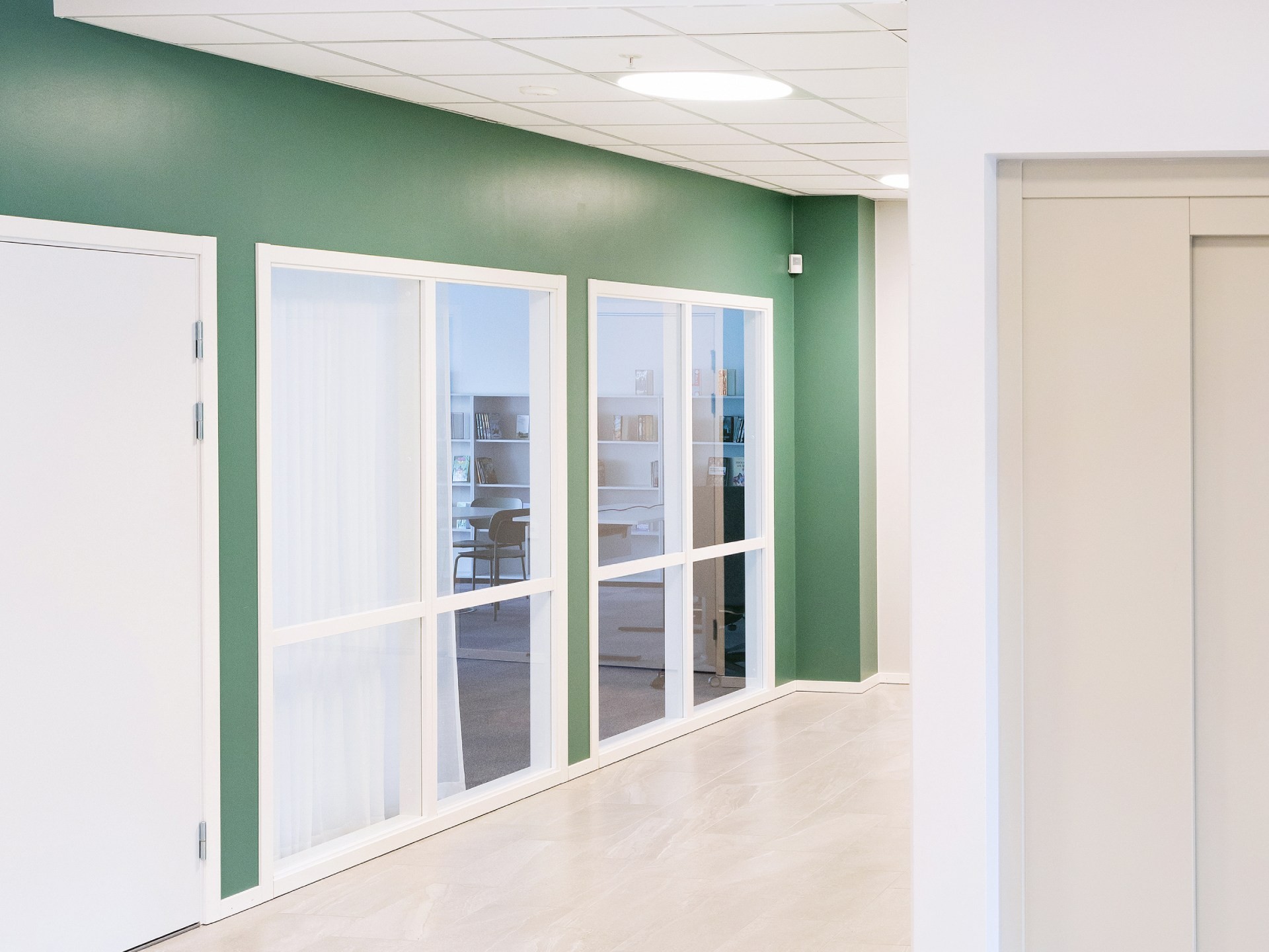 Modern hallway with green walls, white doors, and large windows overlooking an office space.