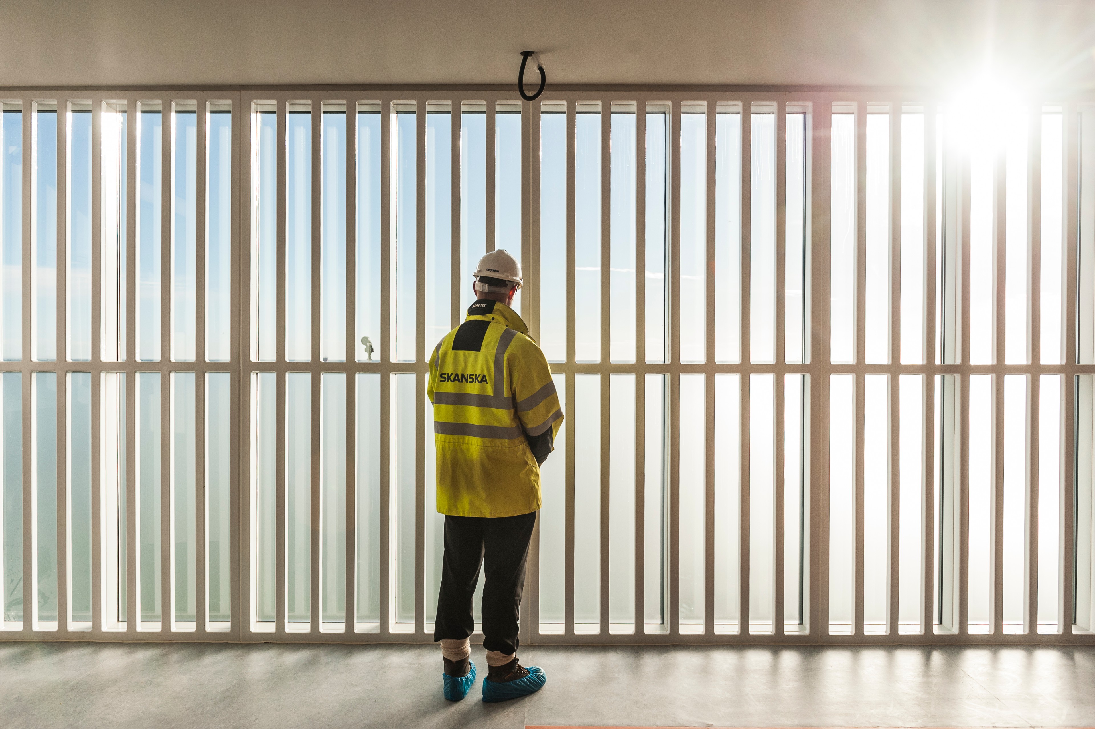 Construction workers in protective equipment observe sunlit site A construction worker in a Skanska jacket and safety helmet stands by large vertical windows, backlit by sunlight.