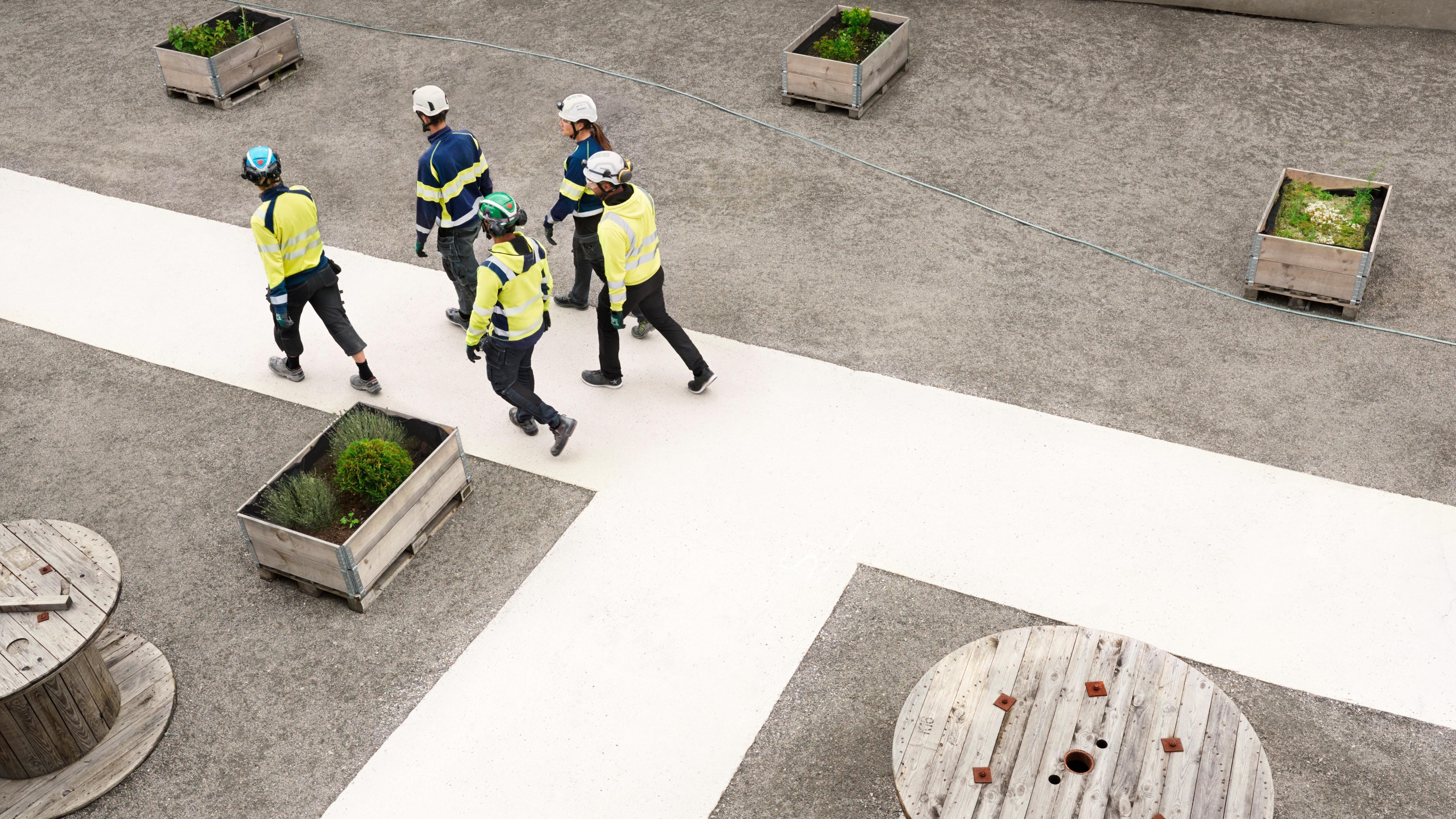 Byggnadsarbetare går på platsen med skyddsutrustning En grupp byggnadsarbetare i skyddsutrustning går på en betongstig omgiven av planteringskärl.