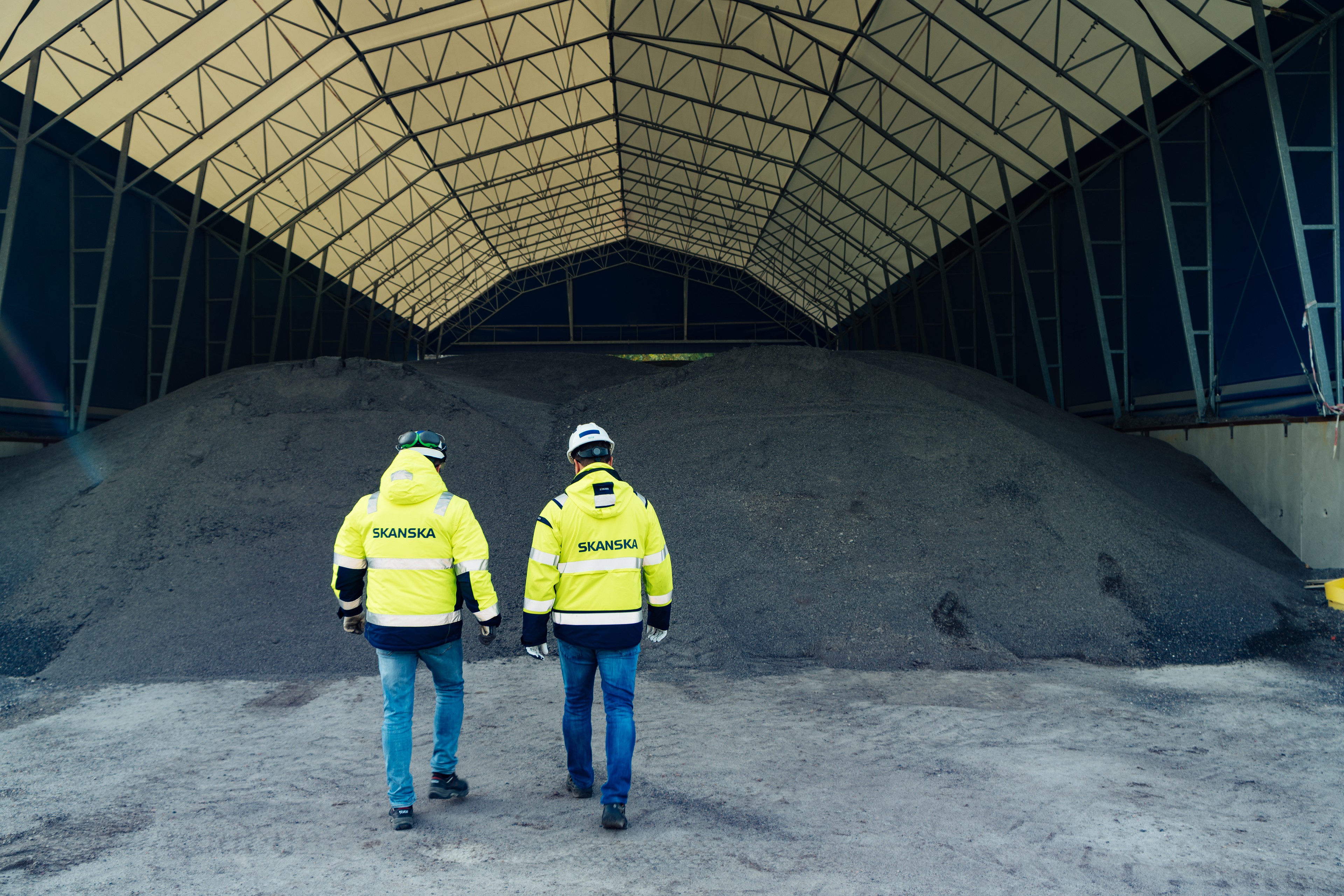 Construction site workers in protective equipment at Skanska Two workers in Skanska jackets walk toward a large gravel pile inside a covered building.