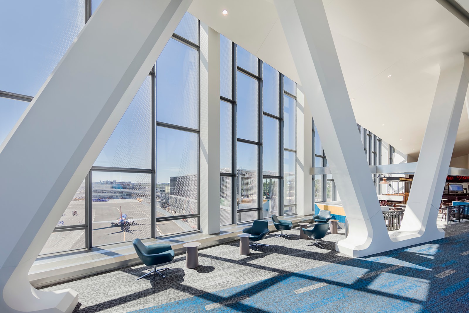 Modern airport lounge with large windows, blue chairs, and a view of the airplanes on the tarmac.