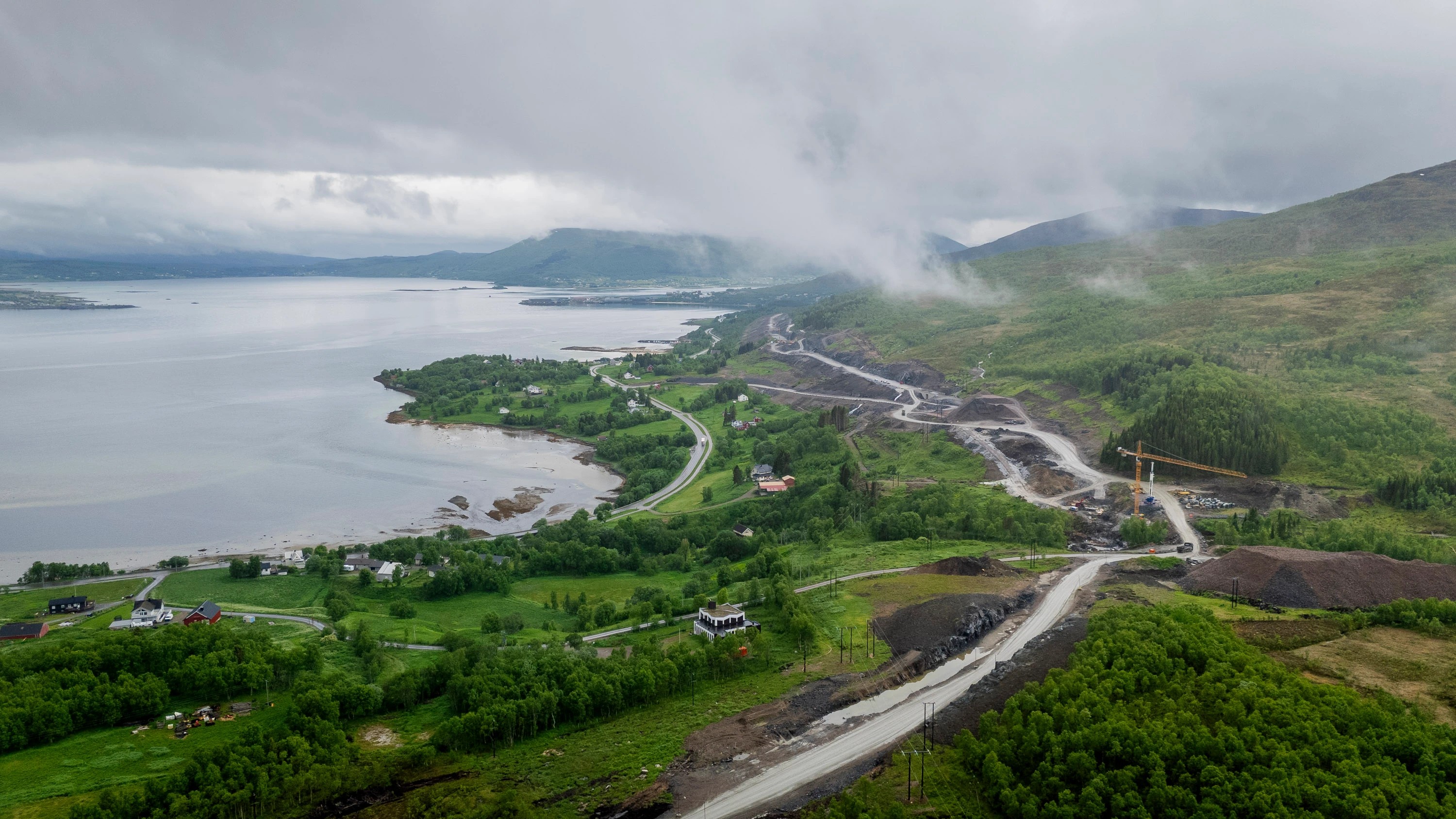 Flygvy av ett kustlandskap med en väg under konstruktion, frodig grönska och molniga himlar.