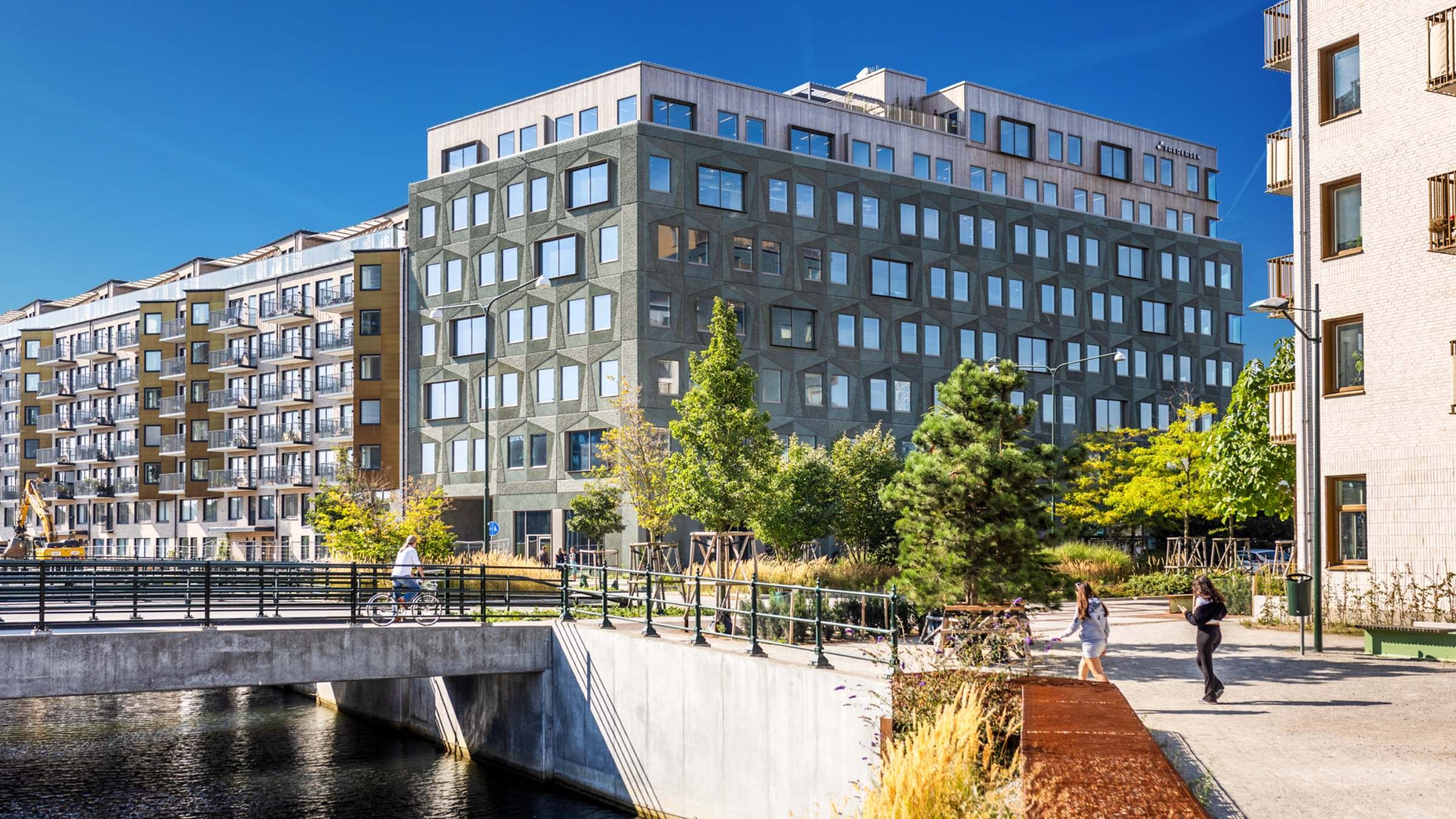 Moderna flerbostadshus vid en kanal med en gångbro och människor som går i närheten.